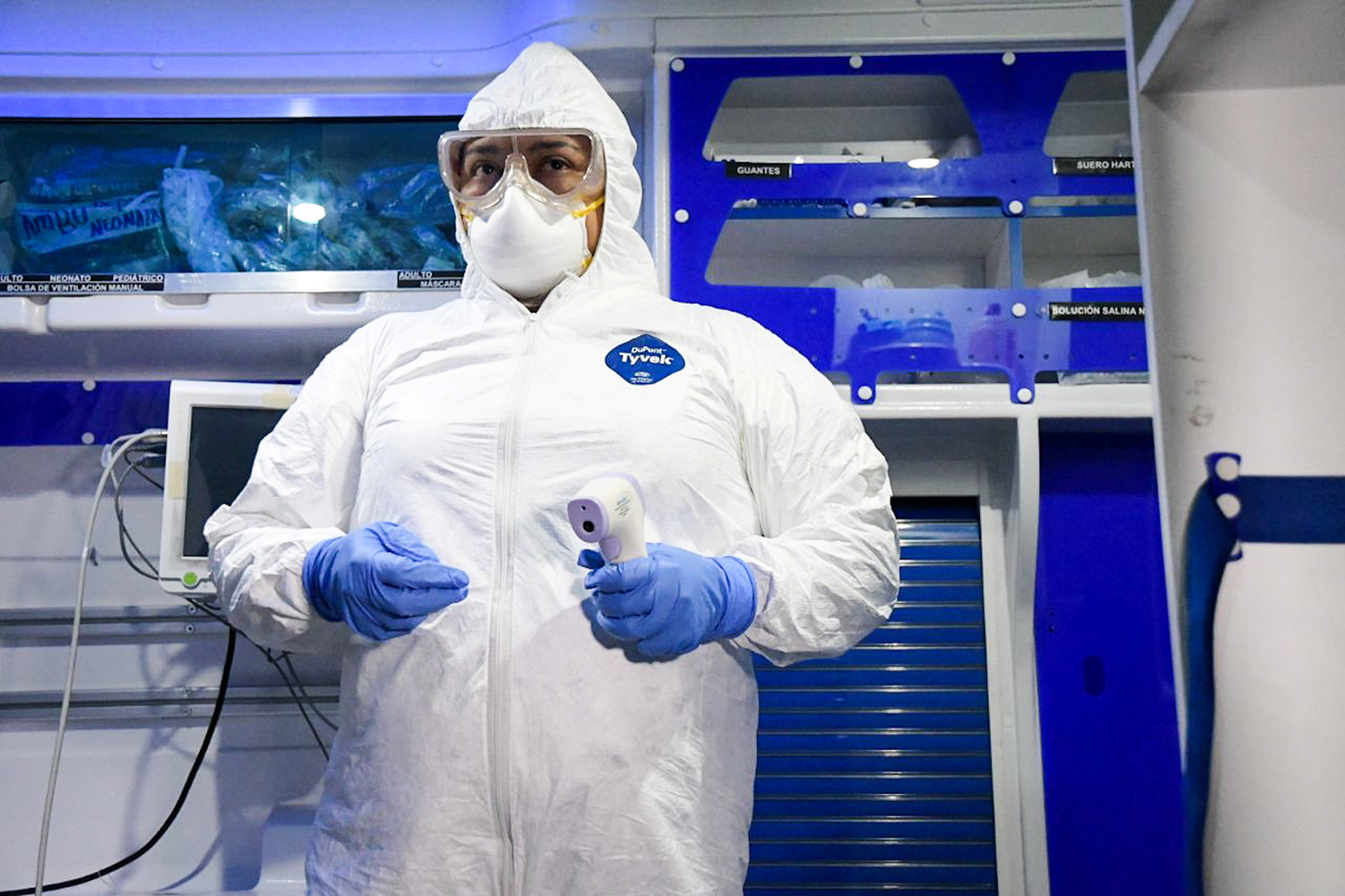 Image shows a worker fully garbed in protective gear head to toe holding a thermometer. Everything in the picture strikingly white or blue.