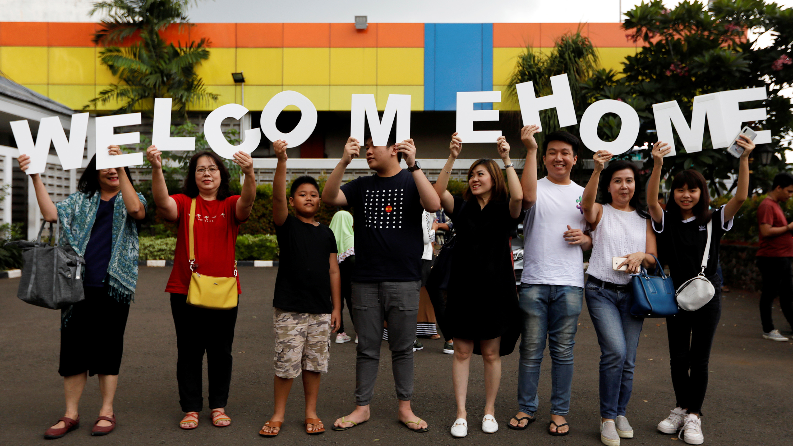 This is a fun photo of a large extended family standing in a parking lot holding a sign that reade welcome home.