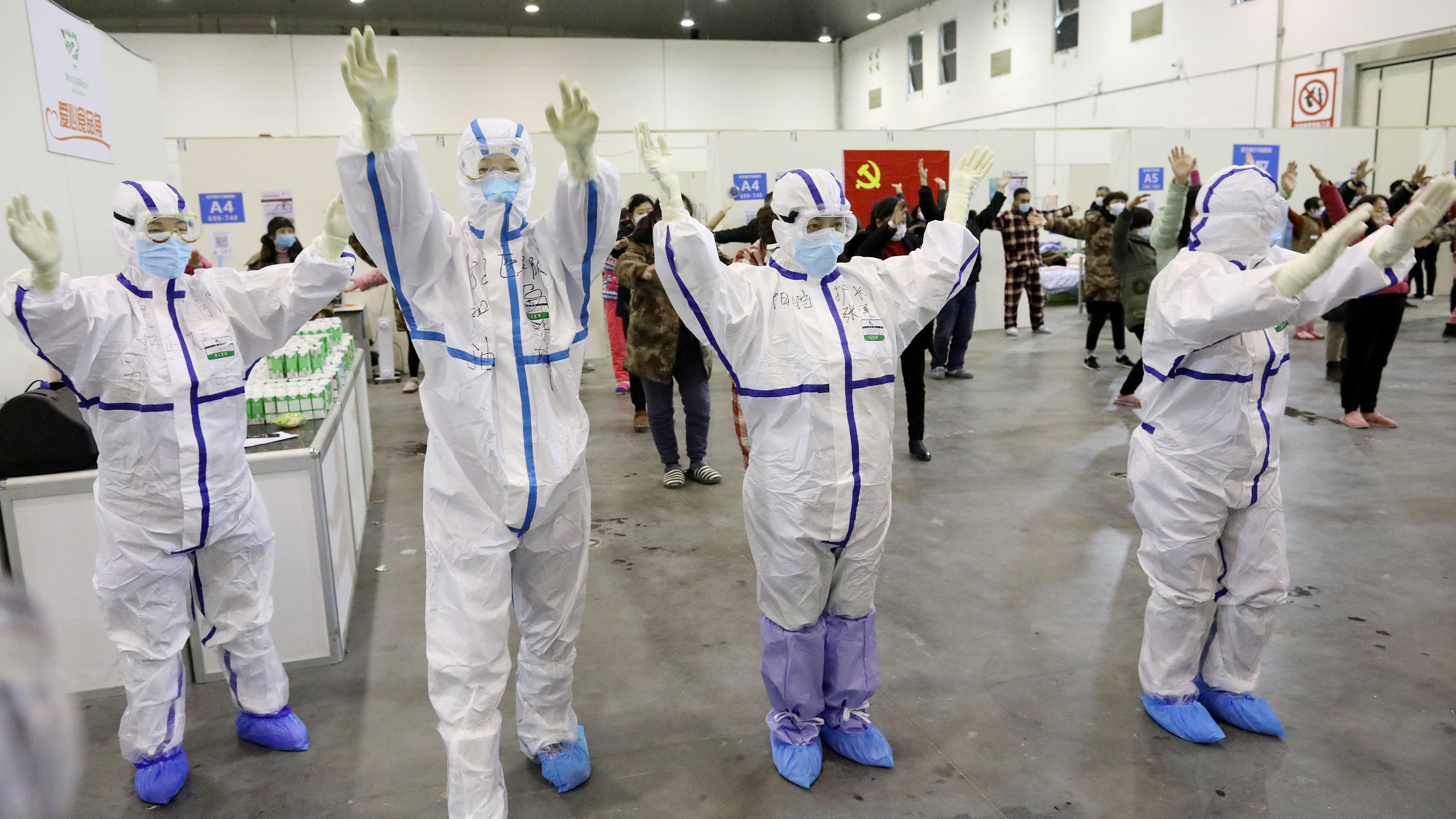 Picture shows a line of health care workers in full protective gear in front of other people who have masks, all with their arms in the air.
