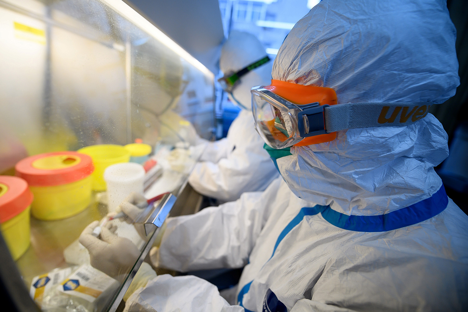 Picture shows two workers in full biohazard protection gear working with specimens under a hood.