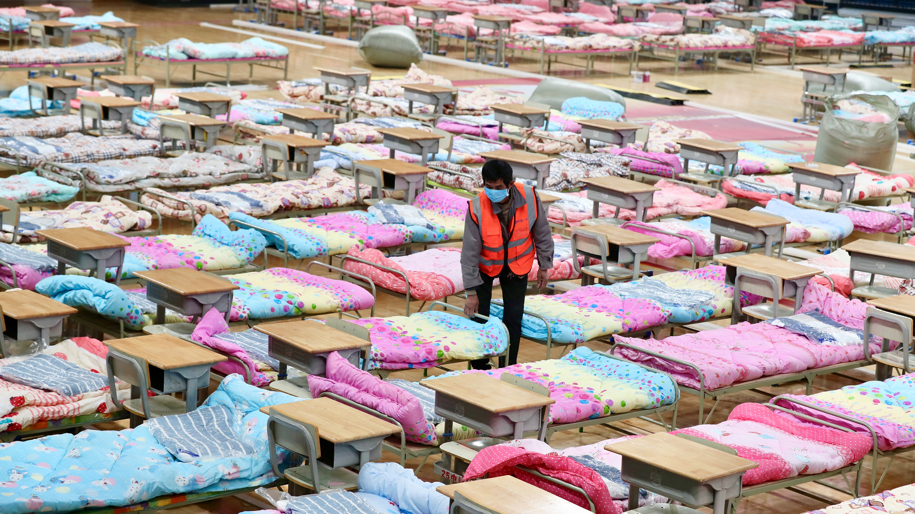 The photo shows a worker adjusting the covers on a bed in a large space filled with hundreds of cot-sized beds covered with white and pink and blue bedding.