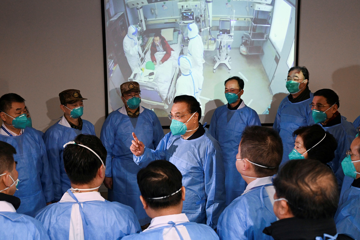 Photo shows a group of men surrounding another man who is speaking and gesturing. They are all wearing protective suits.