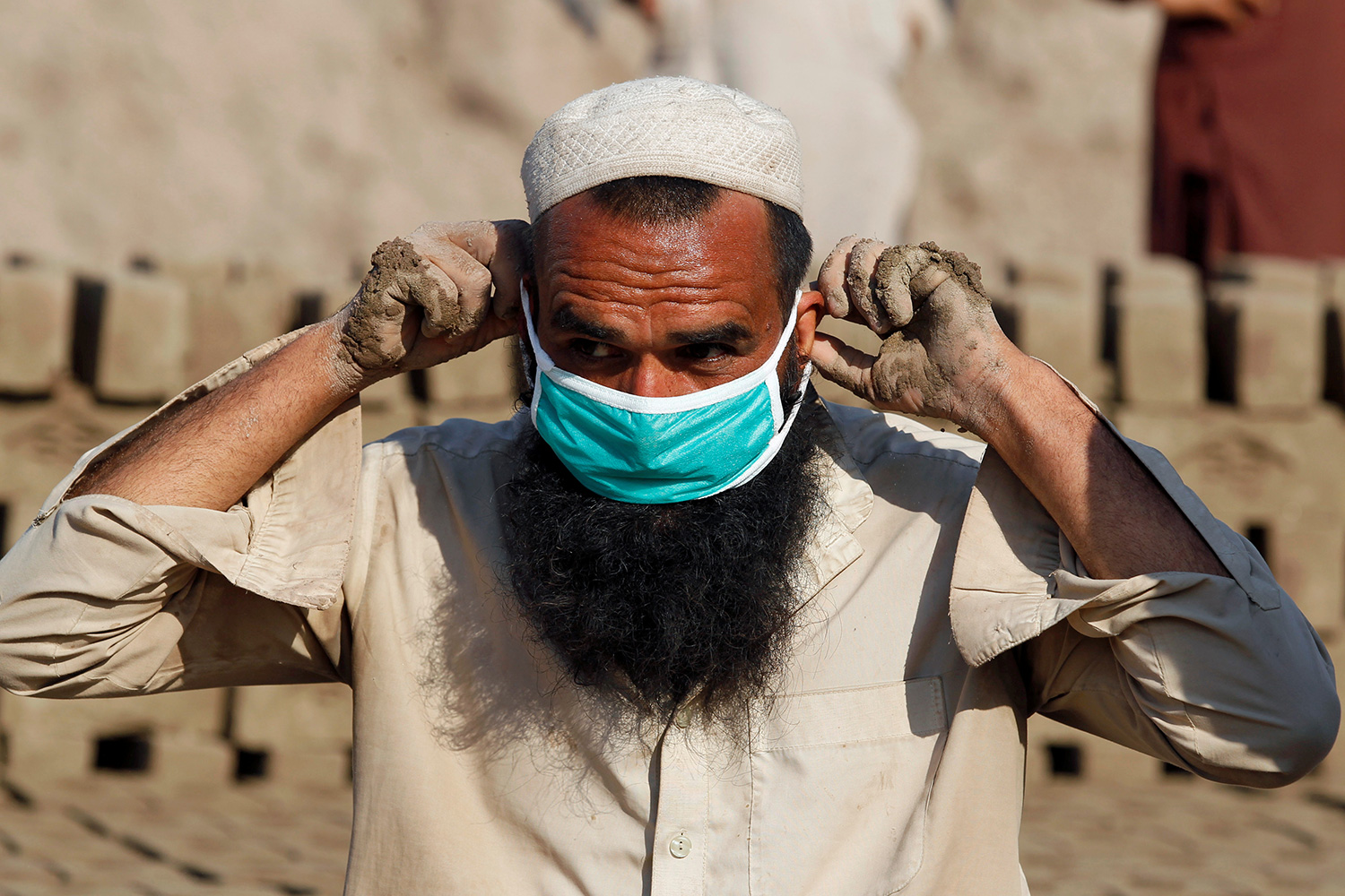 The picture shows a man whose hands are muddy from physical labor donning a blue surgical mask over his face and long beard.