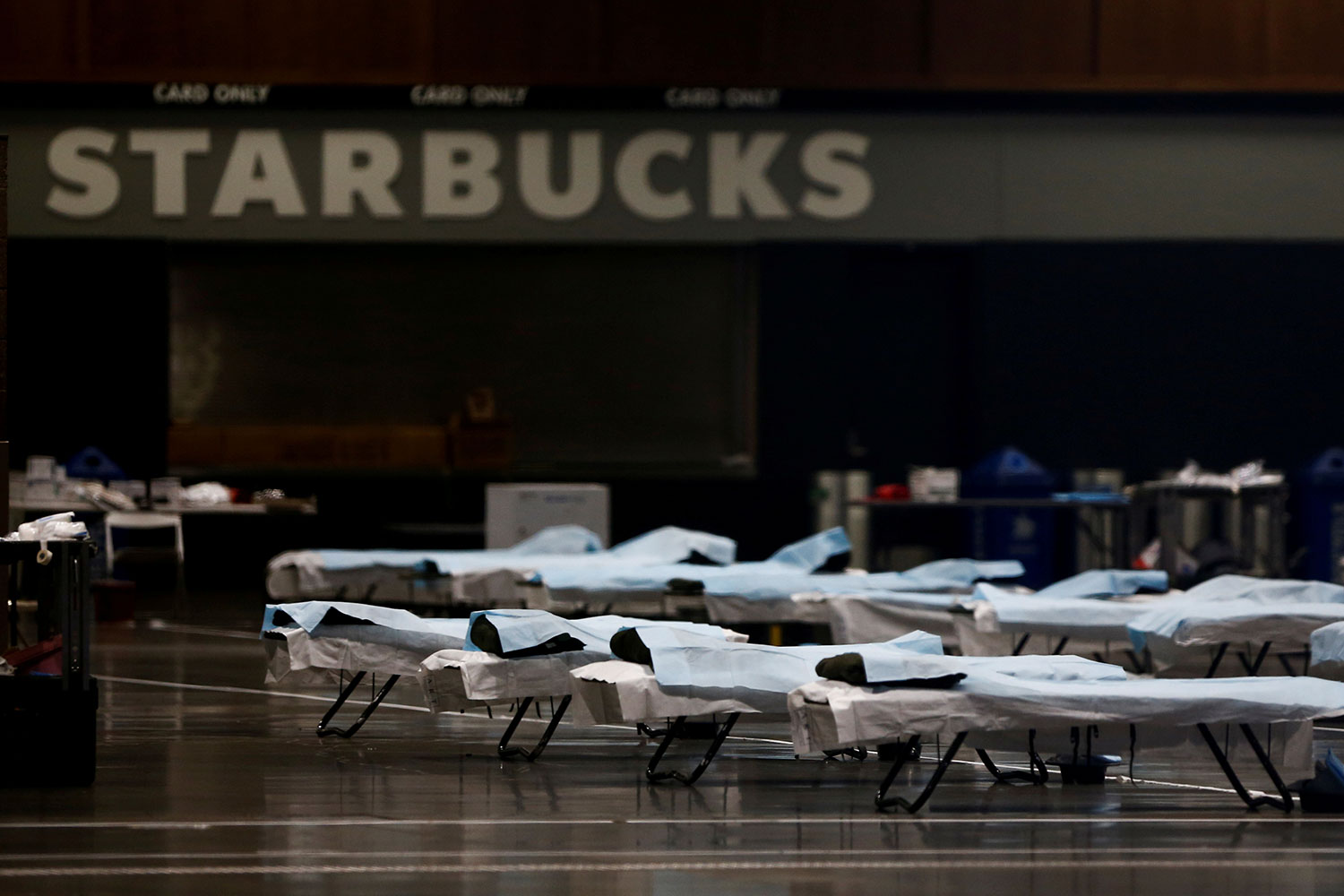 The photo shows a large number of beds lined up in a large open space with a Starbucks sign in the background.