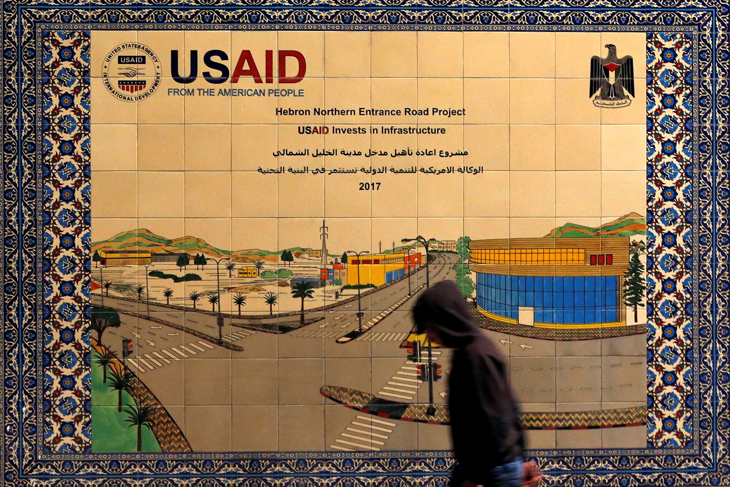 Picture shows a boy silhouetted as he walks past a decorative sign made of fancy tiles that shows a street scene.