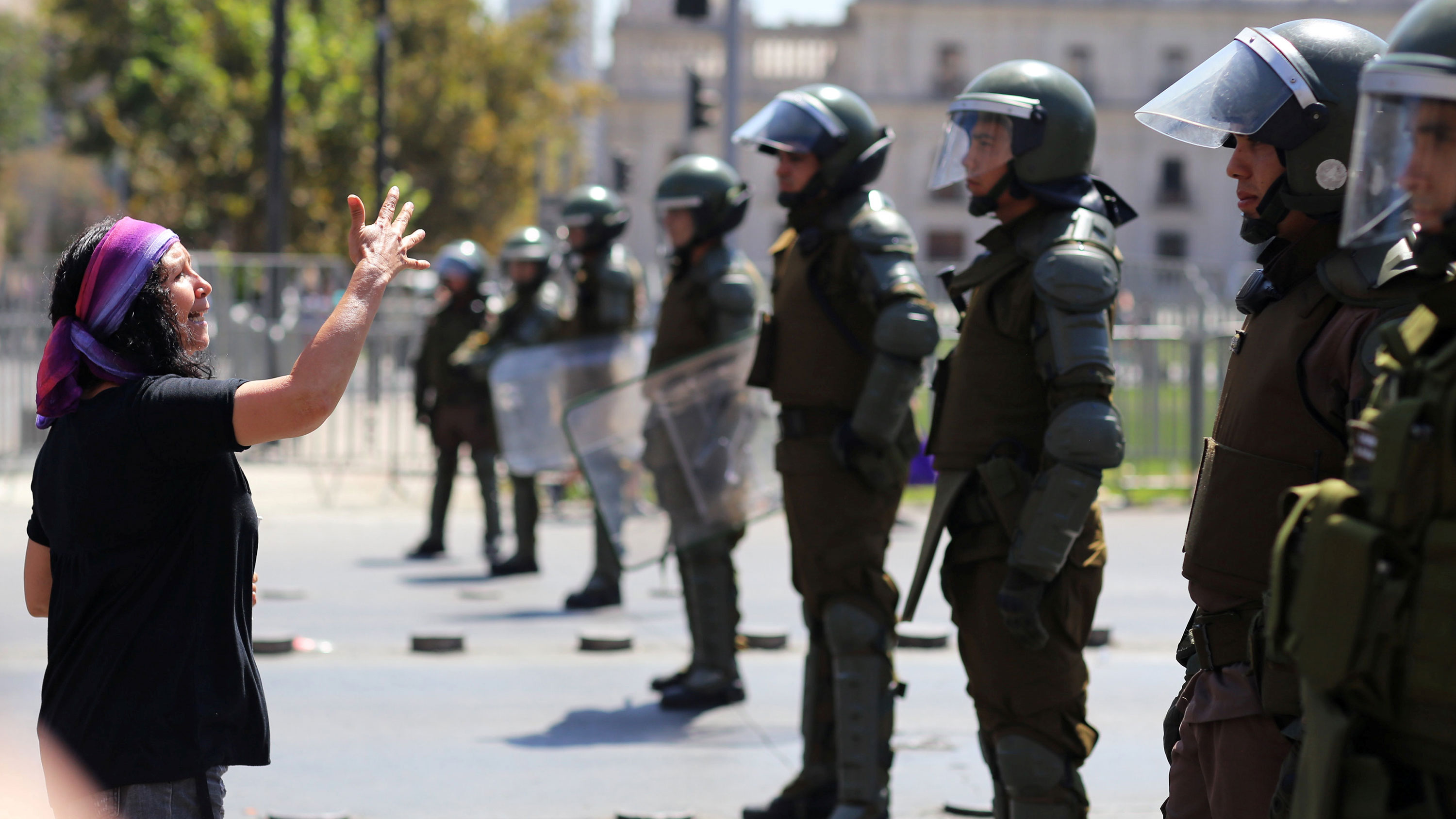 This is a striking photo where a woman wearing a colorful bandana is shouting at a line of police wearing riot gear and gesturing with her hand.