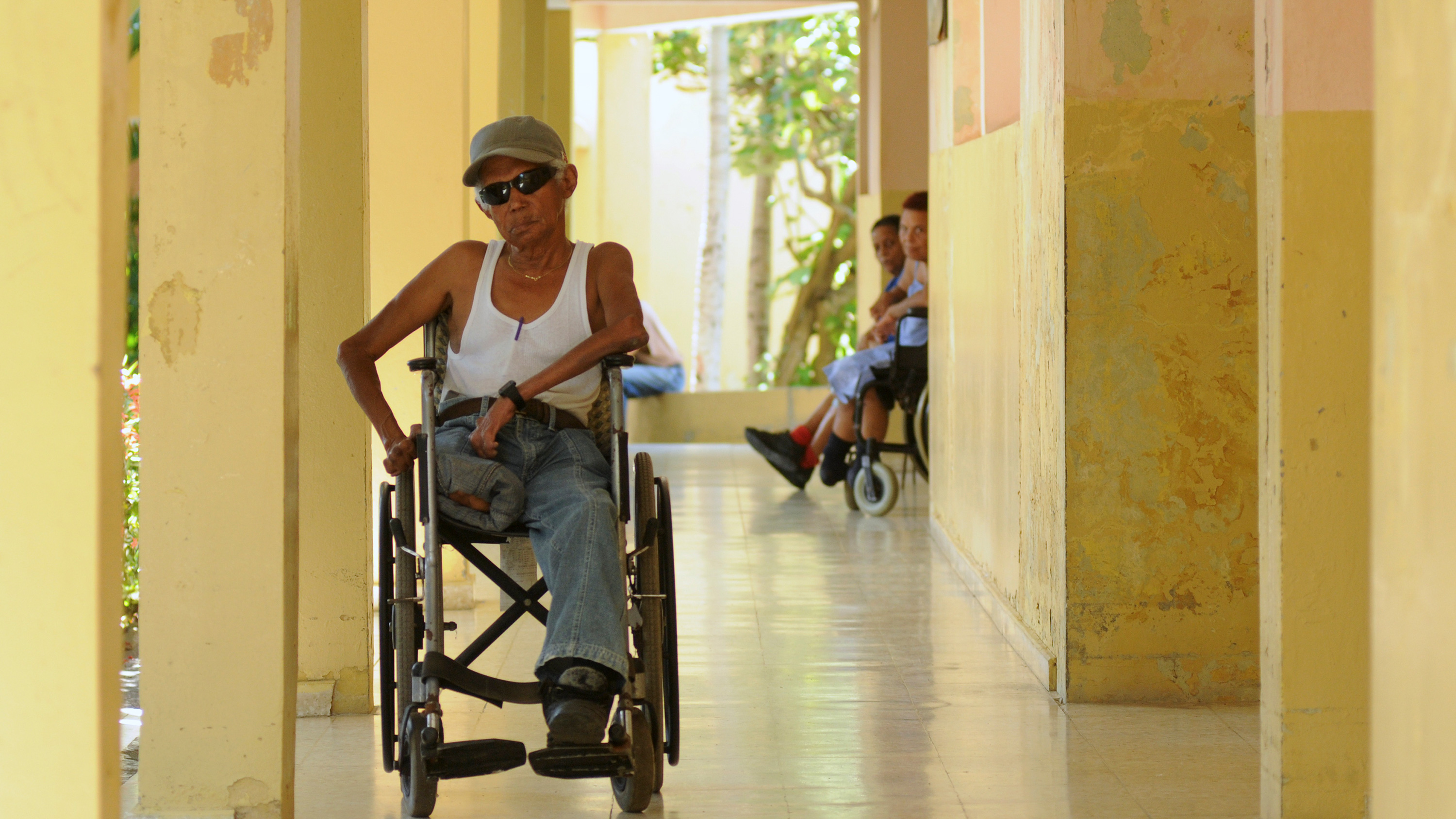 Picture shows Domingo in a wheelchair wearing dark shades and a white tank top. It appears to be a warm summer day.