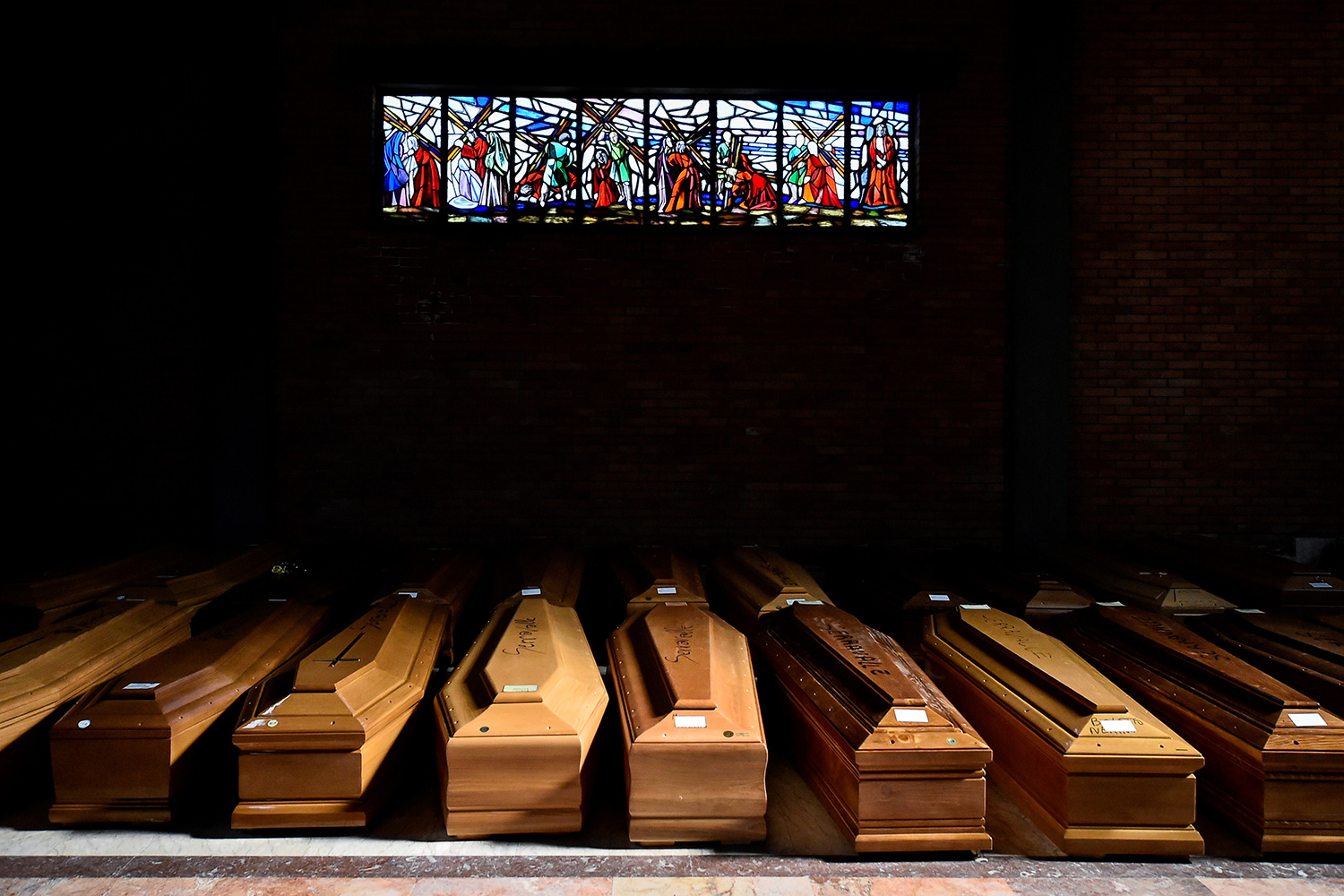 The image shows a huge number of plain coffins in a dimly lit corner of a church beneath a stained glass.