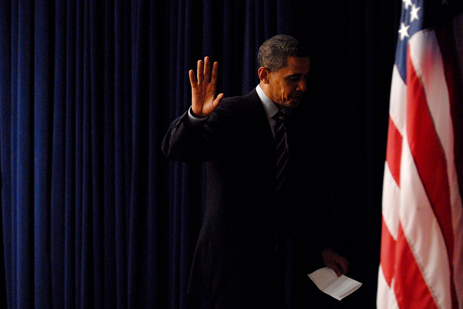 The photo is artistic, with moody, almost too-dark lighting. The former president is holding his hand up as he exits stage left from the podium.