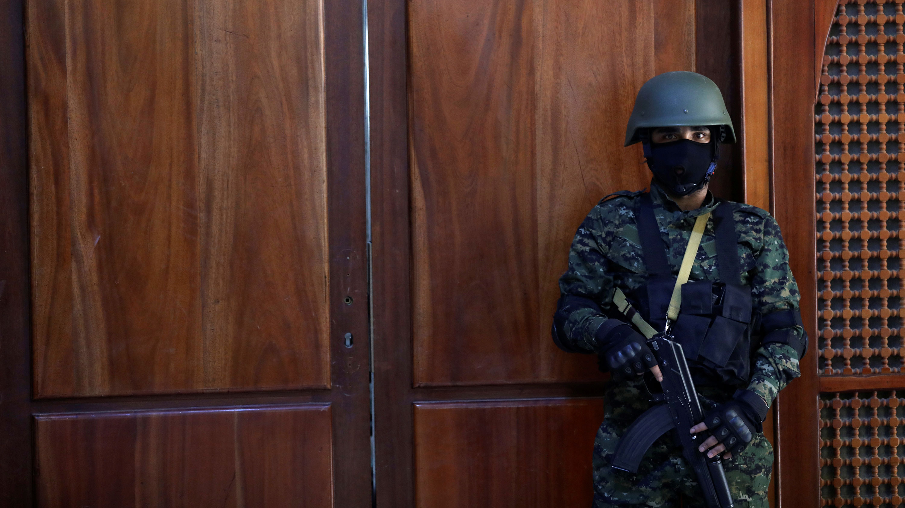 The picture shows a guard with a machine gun standing in front of a wooden door. His mask is black, the door is red.
