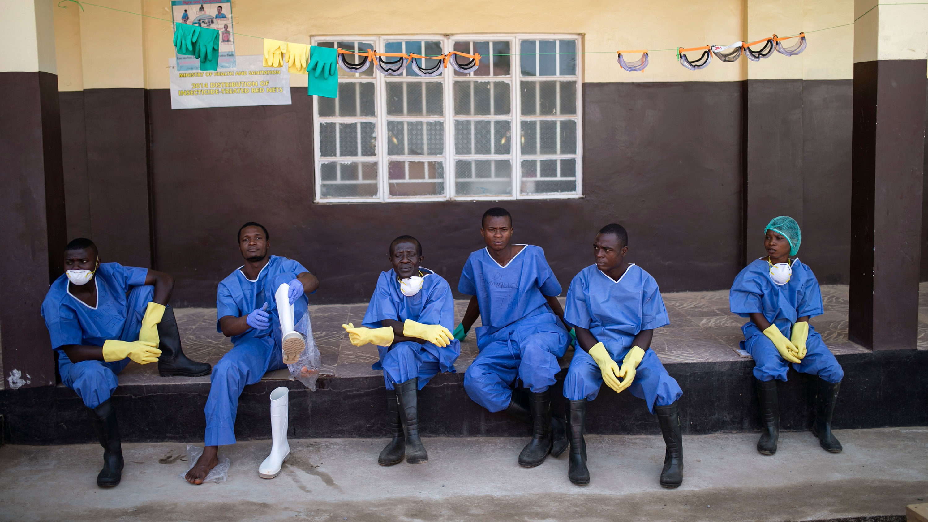 The photo shows six workers on a bench. They are wearing blue scrubs, black boots, and yellow gloves.