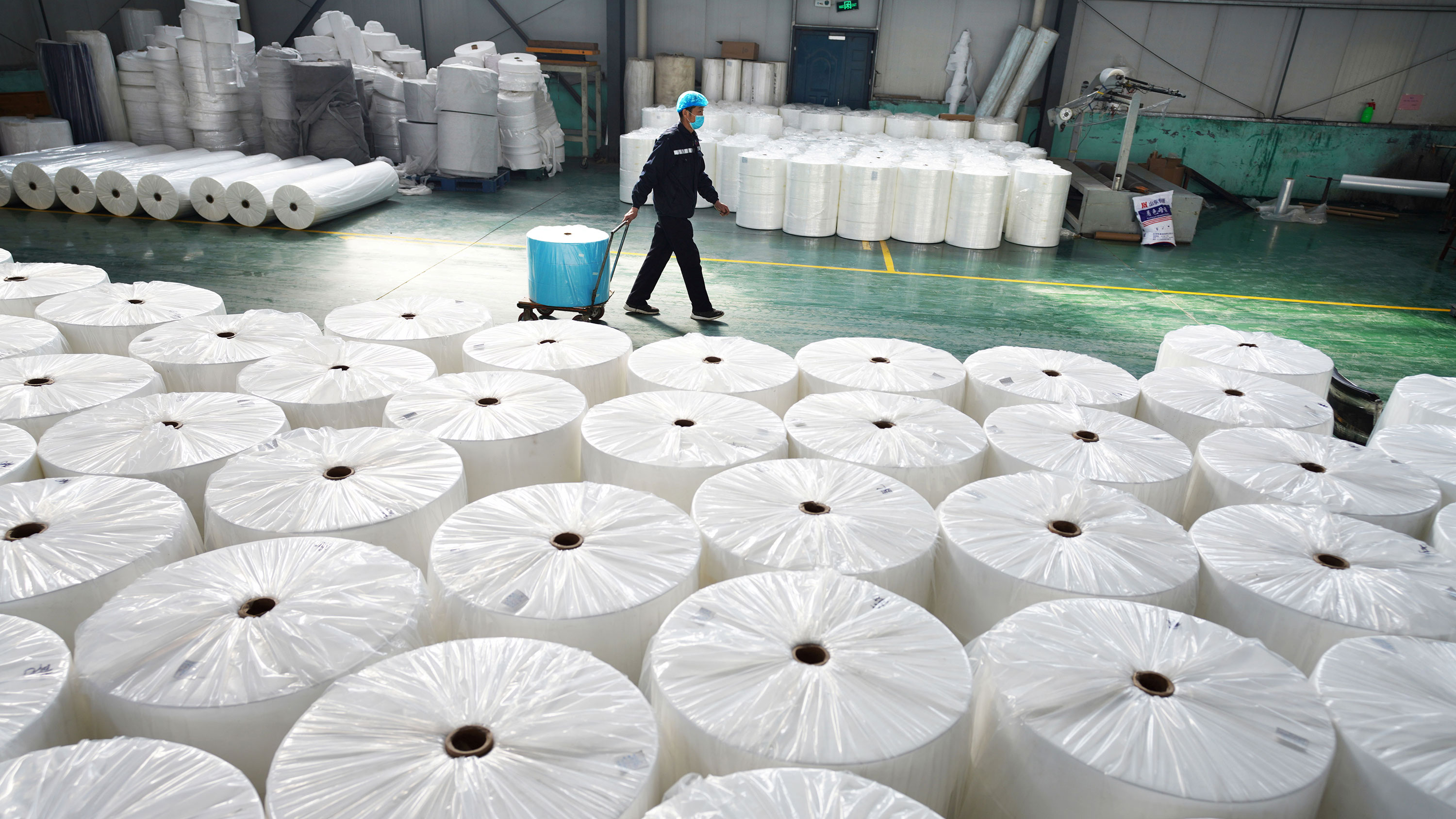 Picture shows a large factory space with a massive stack of large fabric spools in the foreground and a single worker waling by in the background.