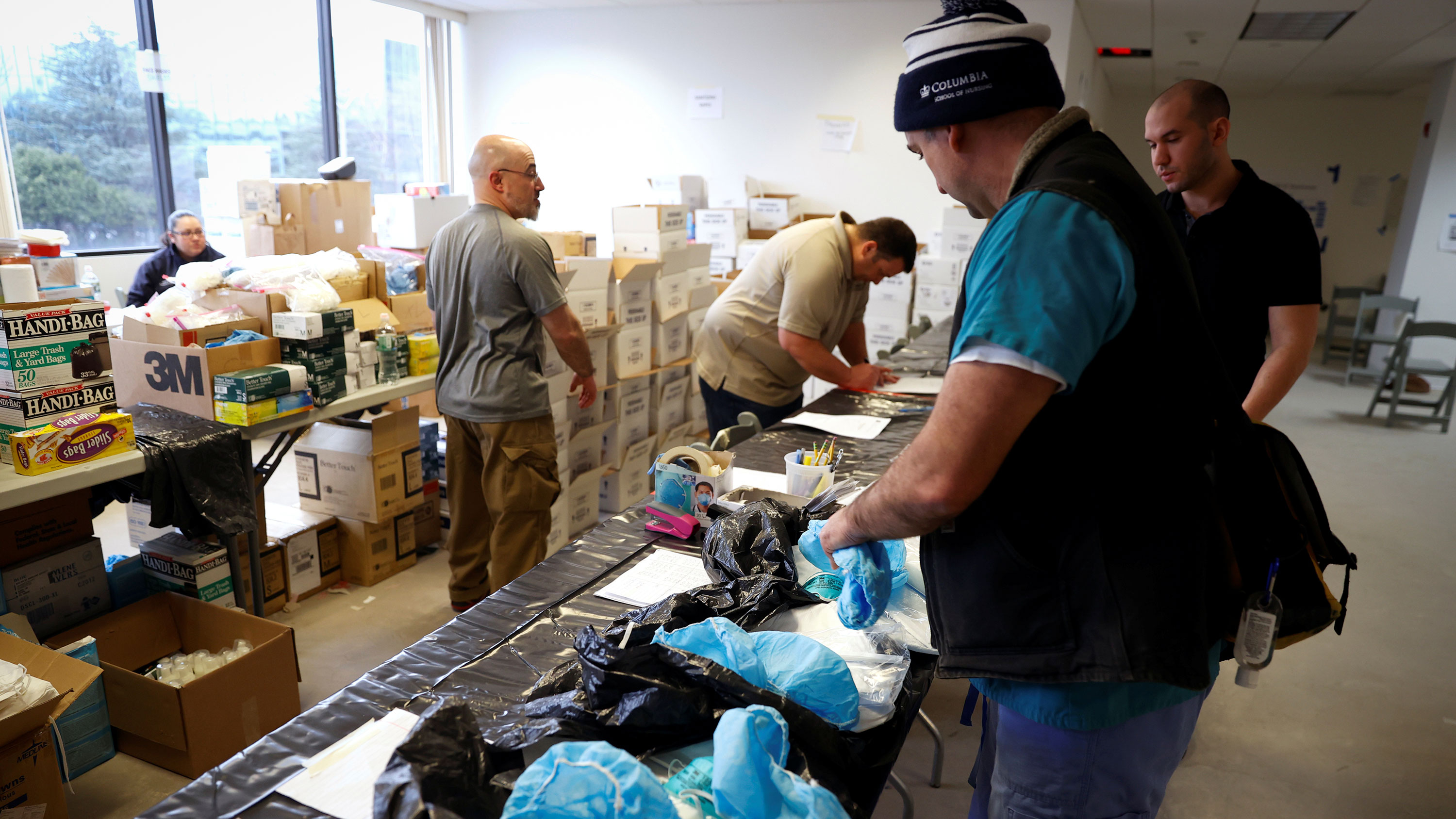 The image shows the nurse in street clothes and a warm cap amid tables heaped with protective gear and several other people busy going through the supplies.
