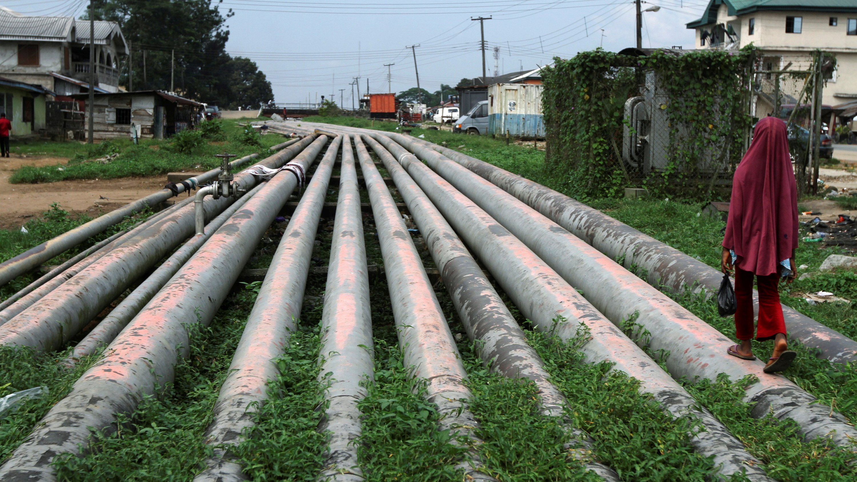 This is a dramatic picture of a dozen or so large pipes, each a couple feet in diameter, running parallel through what appears to be a rural area. The pipes extend off into the distance, forming a natural vanishing point, and a girl wearing red is walking along one of the pipes with her back to the camera.