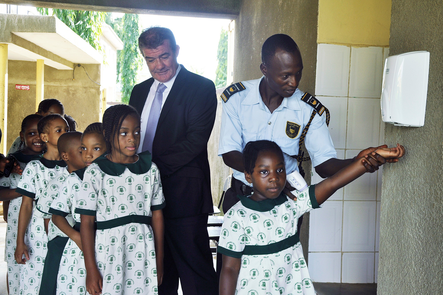 Picture shows a line of girls in school uniforms queueing and being helped at a hand sanitizer station.