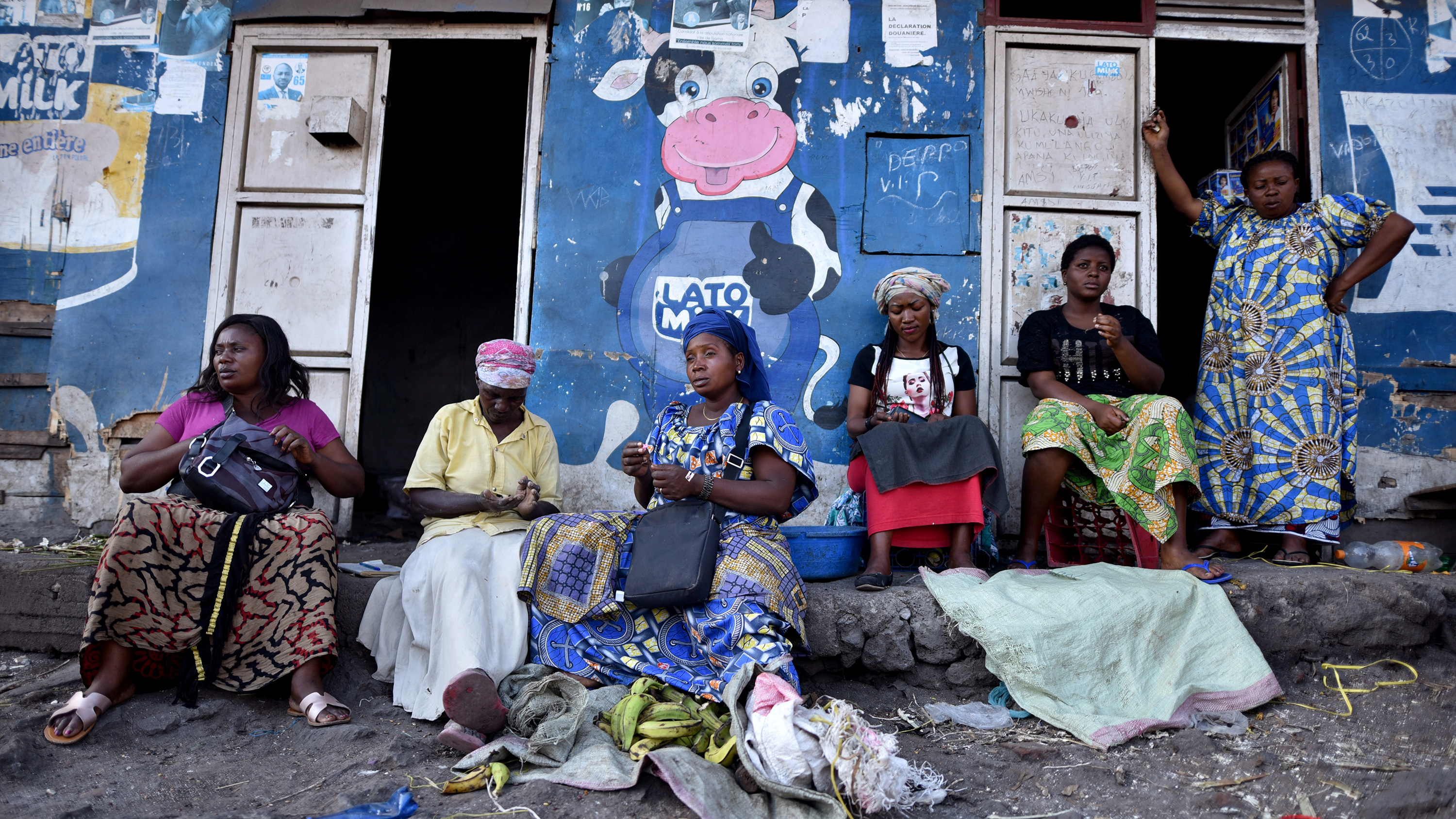 The photo shows a number of women sitting in front of a storefront painted blue and adorned by a large cartoon figure of a cow giving a thumb's up.
