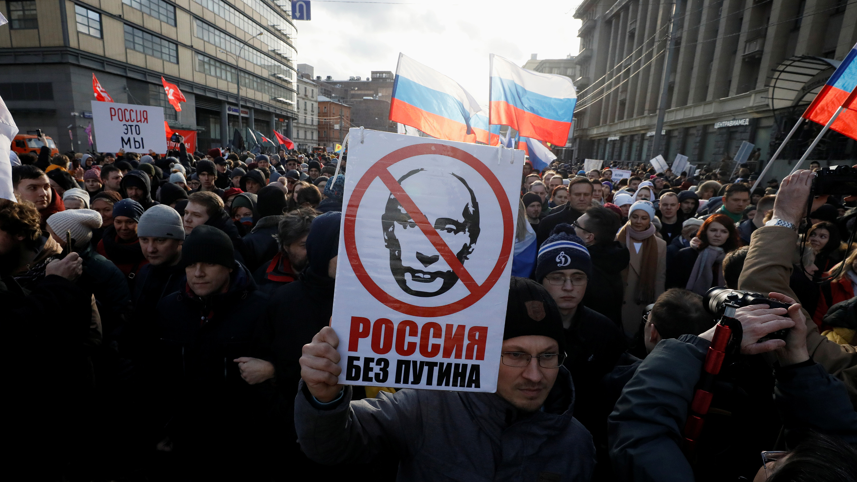 The photo shows a large crowd of marchers. The man in the front holds a placard with a likeness of Vladamir Putin and a red cross over his face. The sign reads: "Russia without Putin."