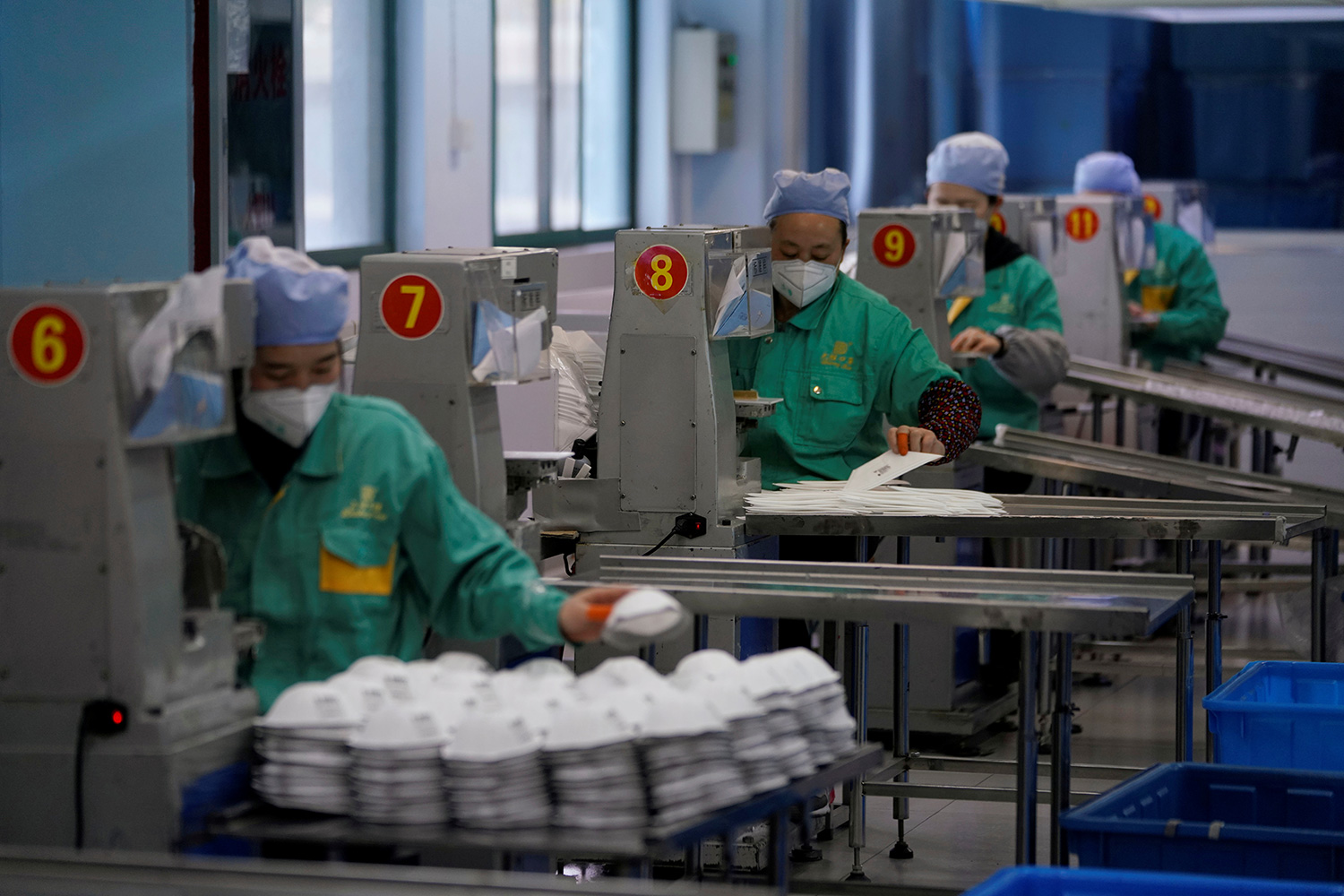 The photo shows a row of workers piling up masks as they roll off machinery. Each one is at a workstation numbered six through twelve.