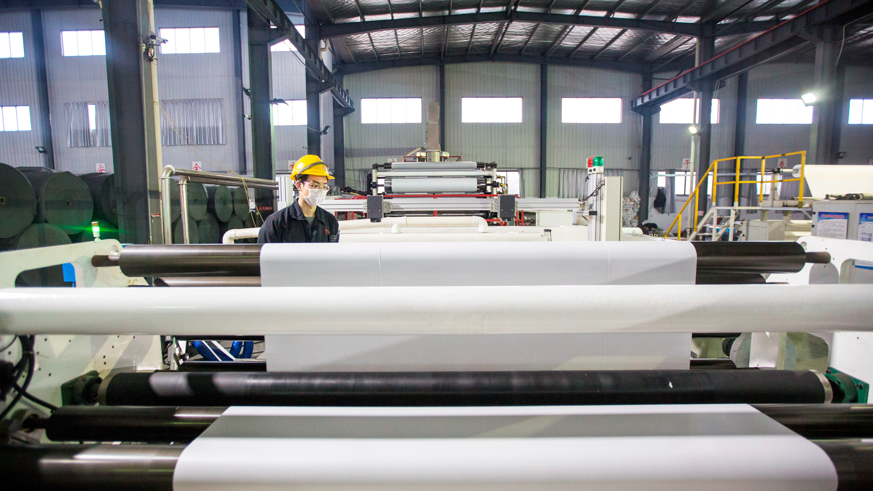 Picture shows a single worker standing amid machinery that has massive rolls of material on spools.