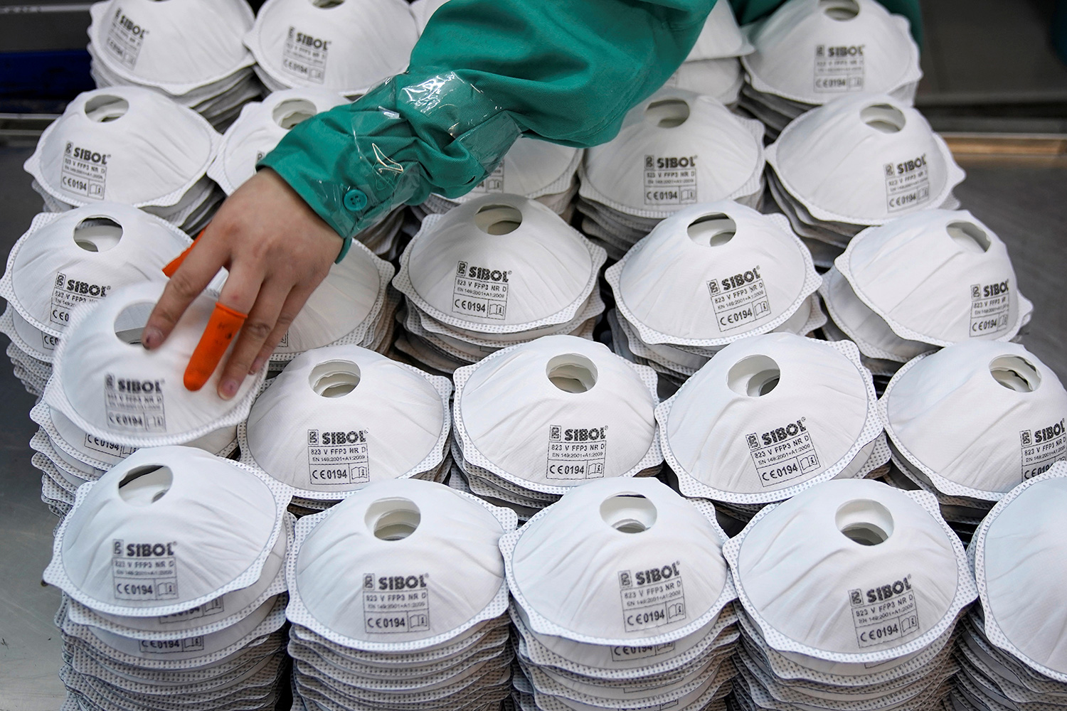 The image shows a human hand with two bright orange bandaids on the thumb and middle finger picking its way across a hige pile of masks.