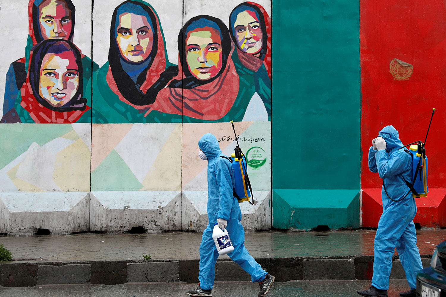 The image shows several workers in blue protective suits walking against a backdrop of a mural painted on the wall with several Afghan women.