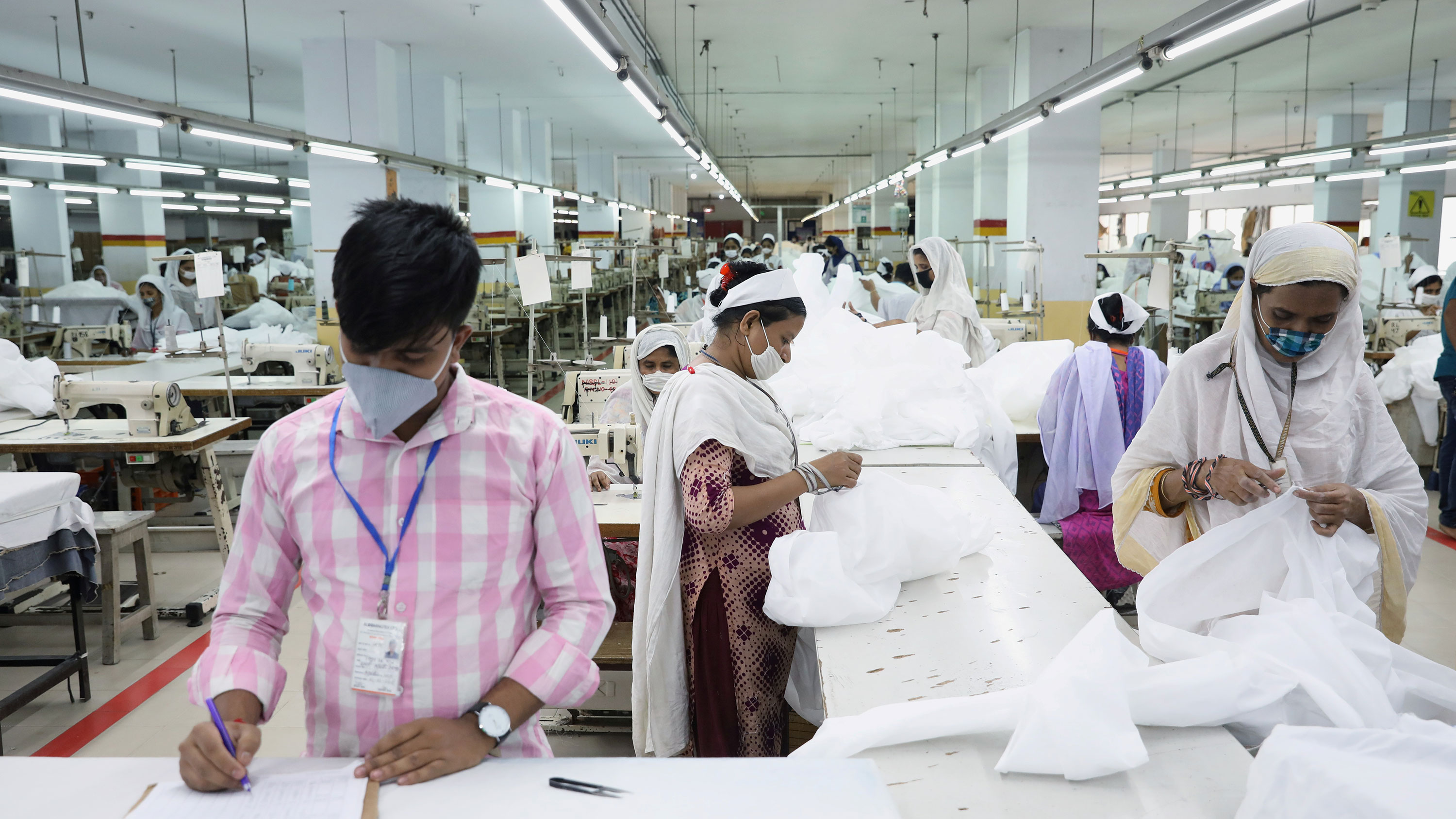 The image shows a factory floor with a man wearing a pink shirt standing in the foreground and several people working—all of whom are wearing masks.