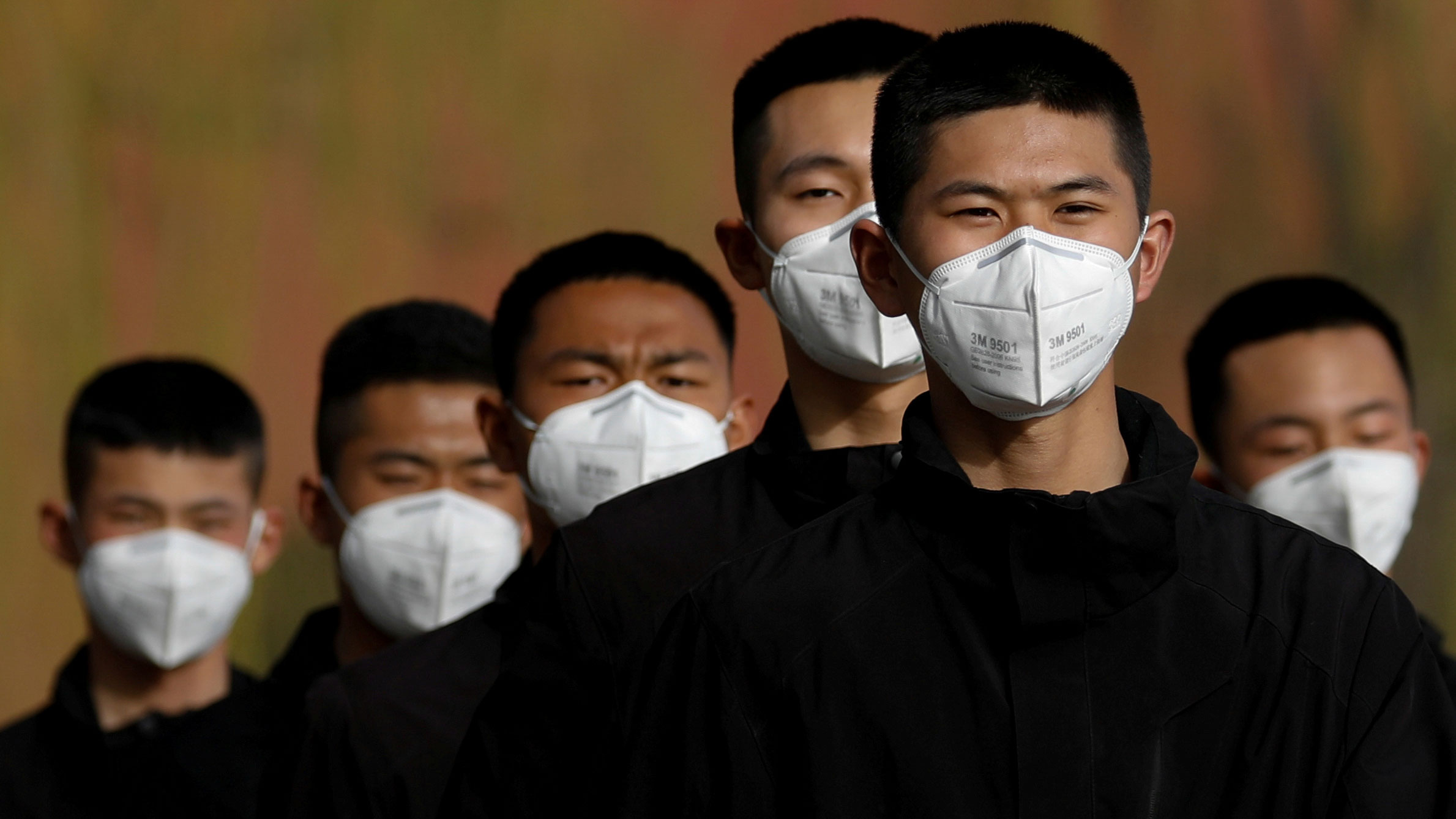 The photo shows half a dozen young men with short cropped hair and all-black clothes in a line, wearing masks.