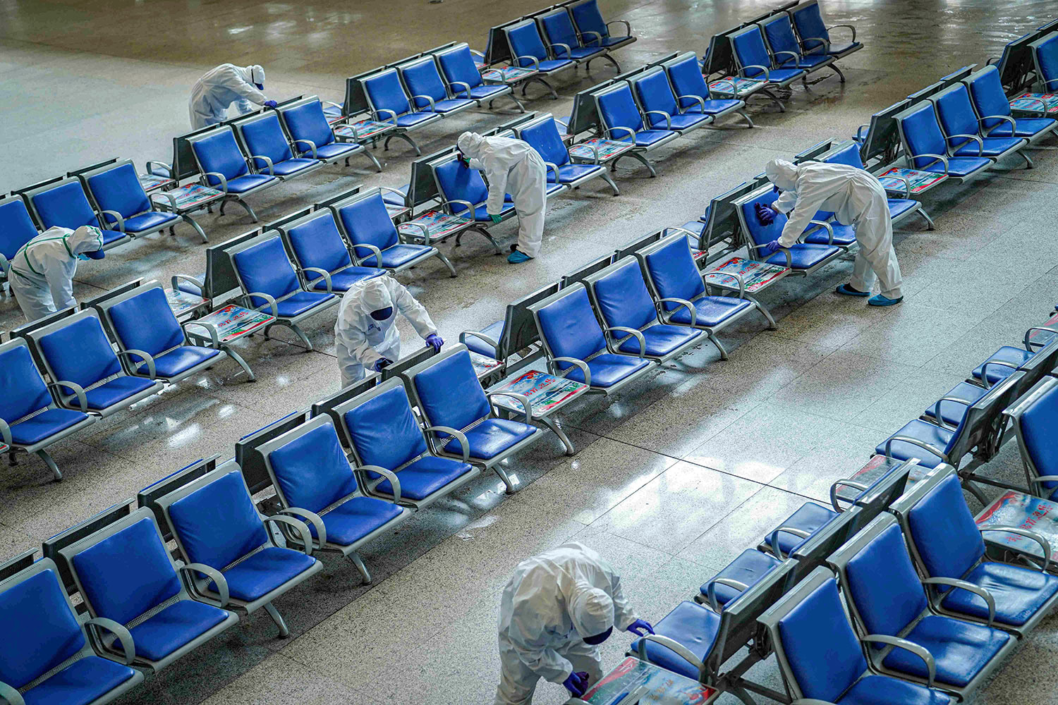The photo shows several long rows of blue benches with workers in protective suits scrubbing them down.
