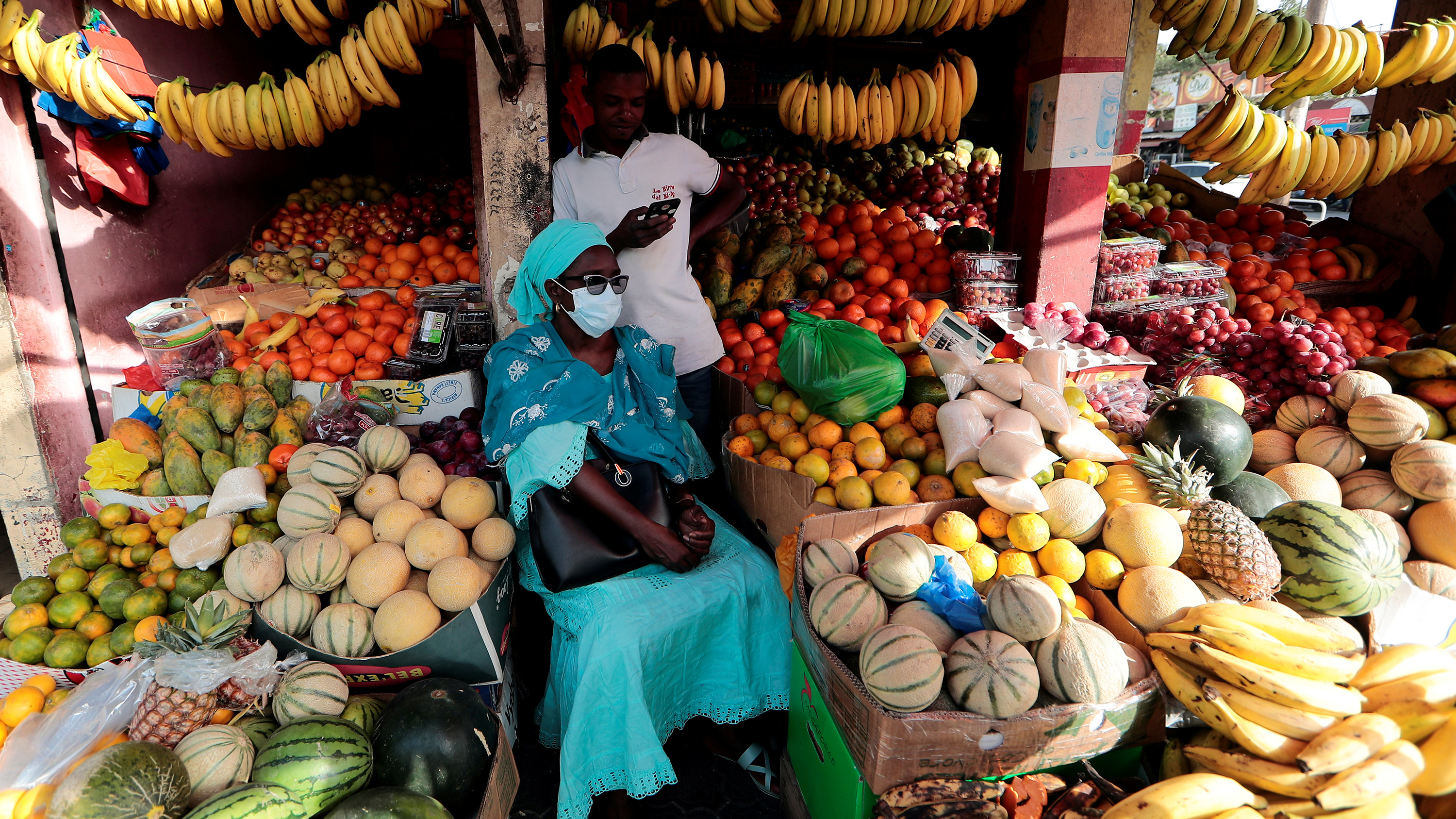 The photo shows a woman wearing a mask sitting amid an abundance of fruit in a tiny stand.