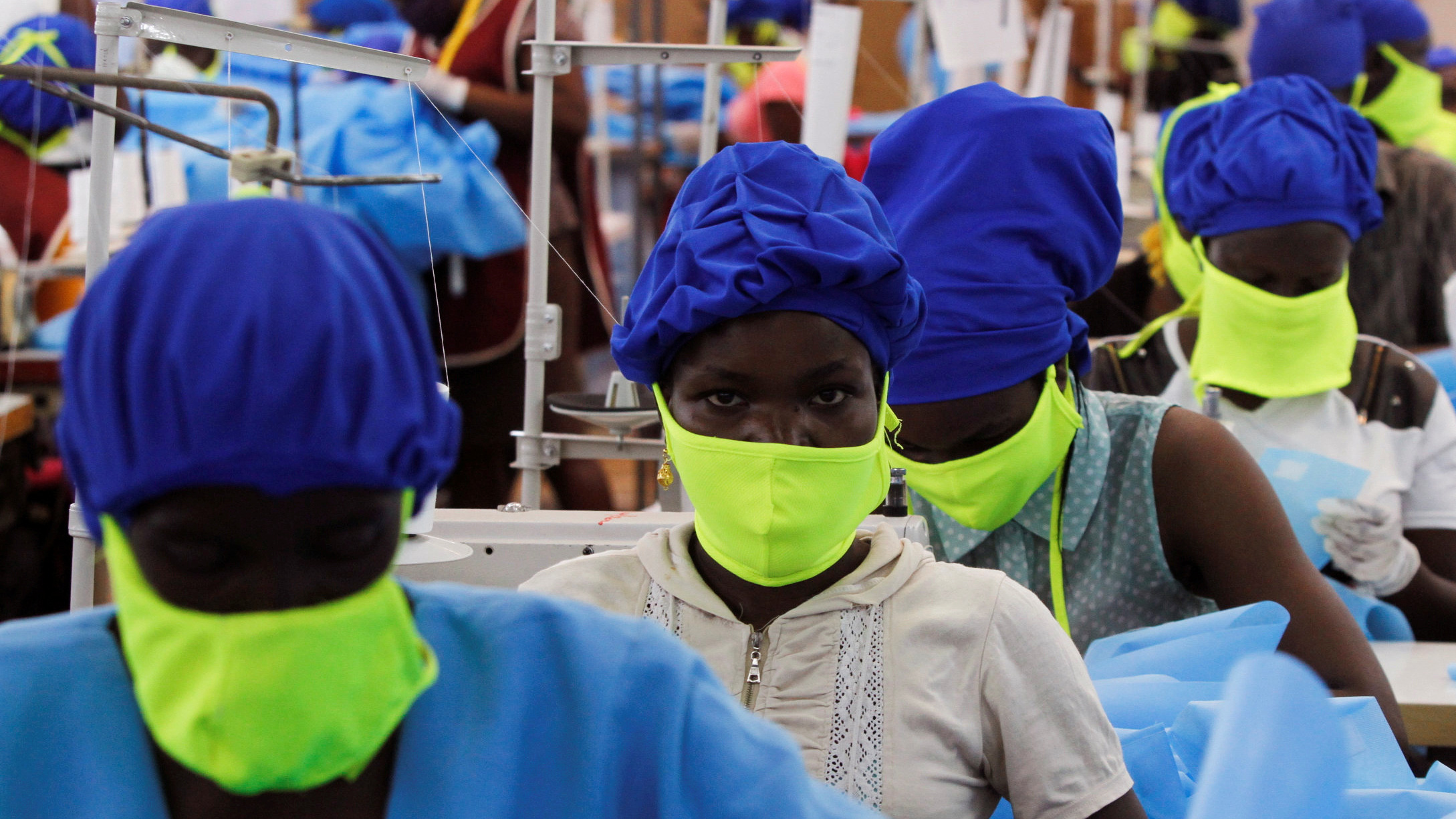 The photo shows a row of workers wearing protective blue suits and bright green facemasks working in a factory.