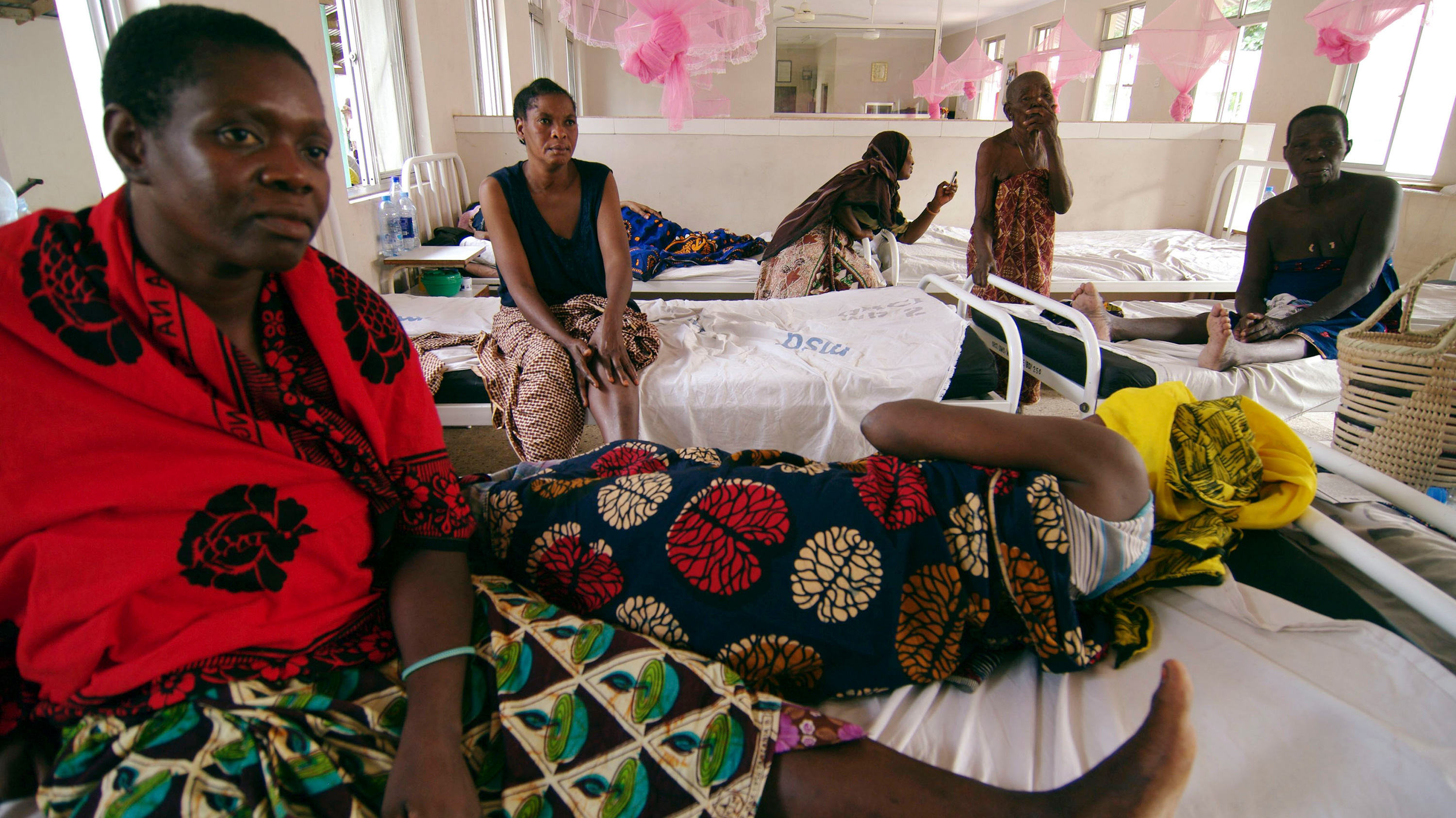 Picture shows Kondogoza in a brightly-colored wrap sitting up in bed while her bedmate stretches out next to her. Several other women can be seen in the background on other beds.