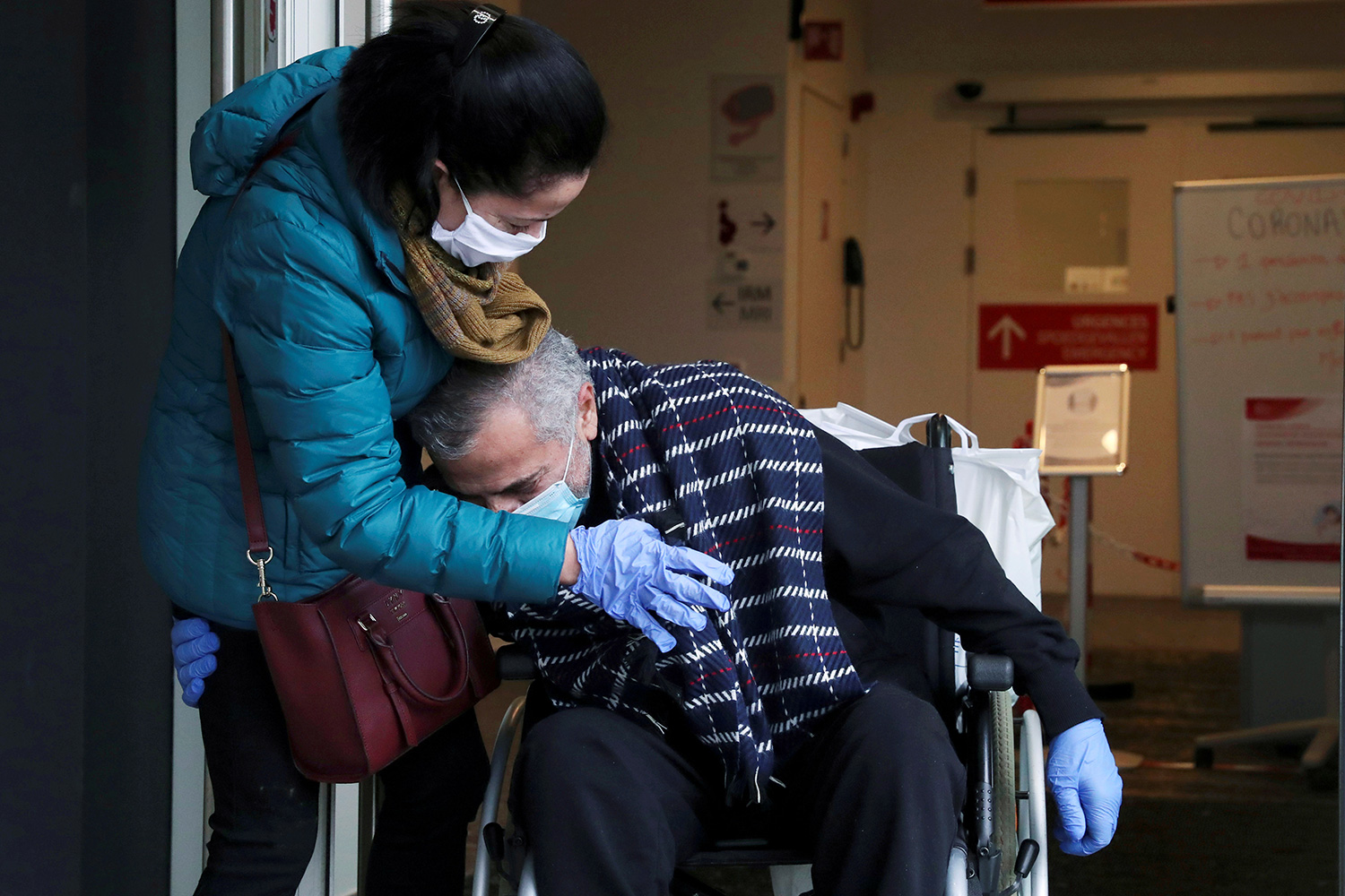 Image shows the doctor in a wheelchair hugging at his wife's torso as she leans into him. They are both wearing masks and gloves.