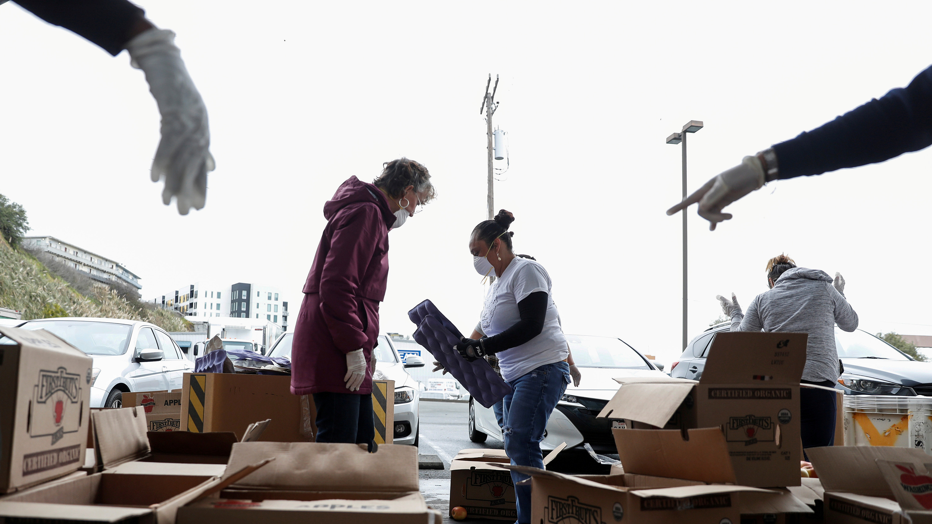 The photo shows a couple volunteers among boxes and boxes of apples with a few arms pointing in each corner of the frame from people who are standing off-camera.