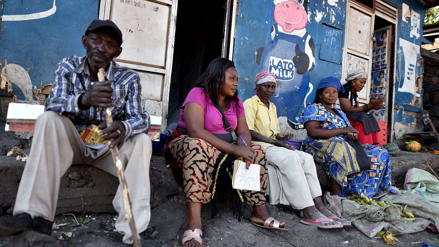 The photo shows a number of women and one man sitting in front of a storefront painted blue and adorned by a large cartoon figure of a cow giving a thumb's up.