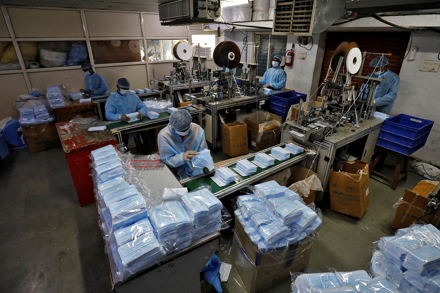 The photo shows a factory interior where several workers are at stations stacked high with blue surgical-style masks.