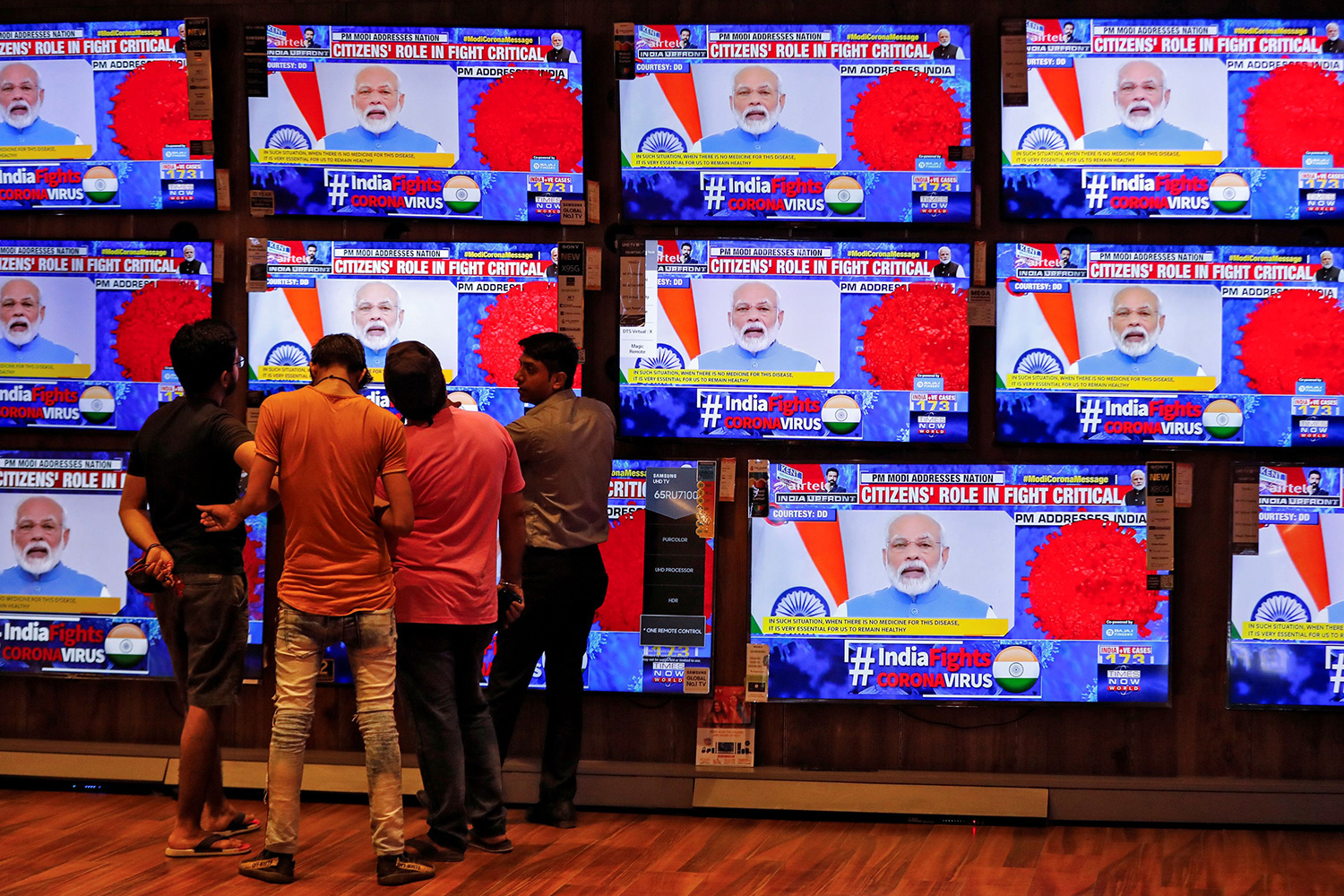 The photo shows a few people standing in front o a showroom with a large stack of TV monitors and the prime minister on all of them giving a speech to the nation.
