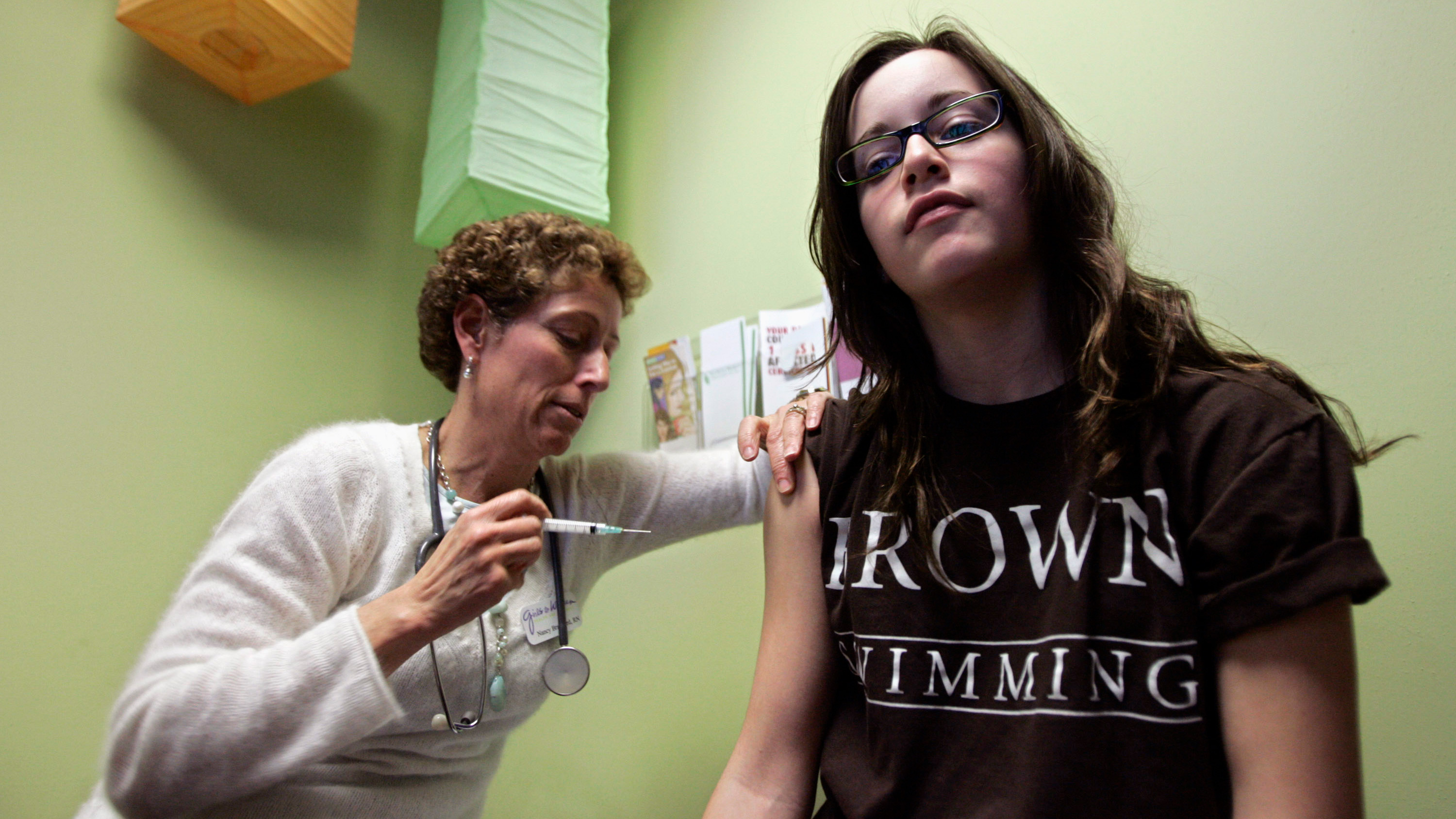 The photo shows a nurse giving a shot to a teemage girl who is looking straight at the camera.
