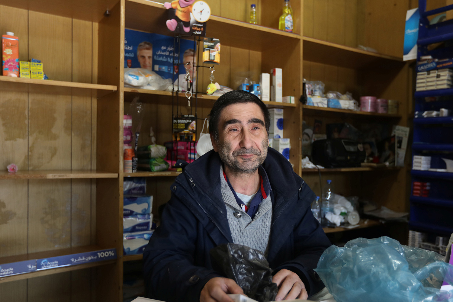 Picture shows an older man with a kind face smiling at the camera from behind a counter in front of an array of empty shelves.