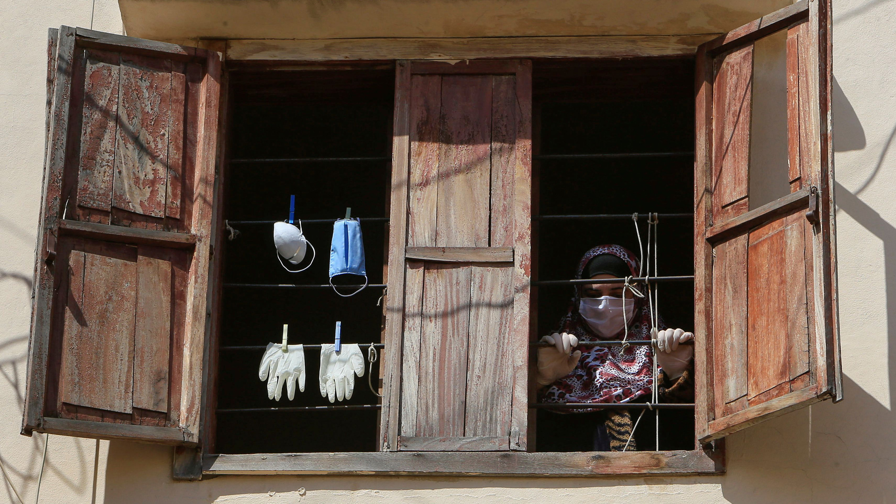 The photo shows a pair of old wooden shutters next to a pair of open windows in an old building with the sun beating don on it. Seen hanging in one window is a pair of gloves and a pair of masks. A woman can be seen looking out from the dark interior of the room within.