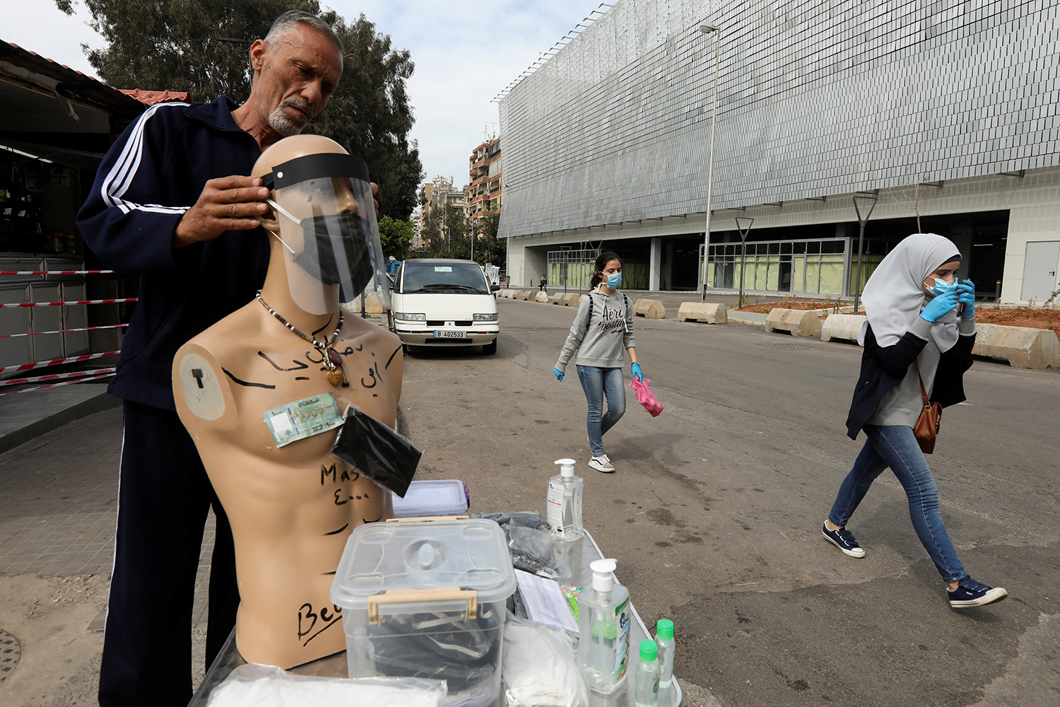 The photo shows the man fitting a clear plastic mask on a manikin next to a makeshift huckster table on a busy street with people walking by.