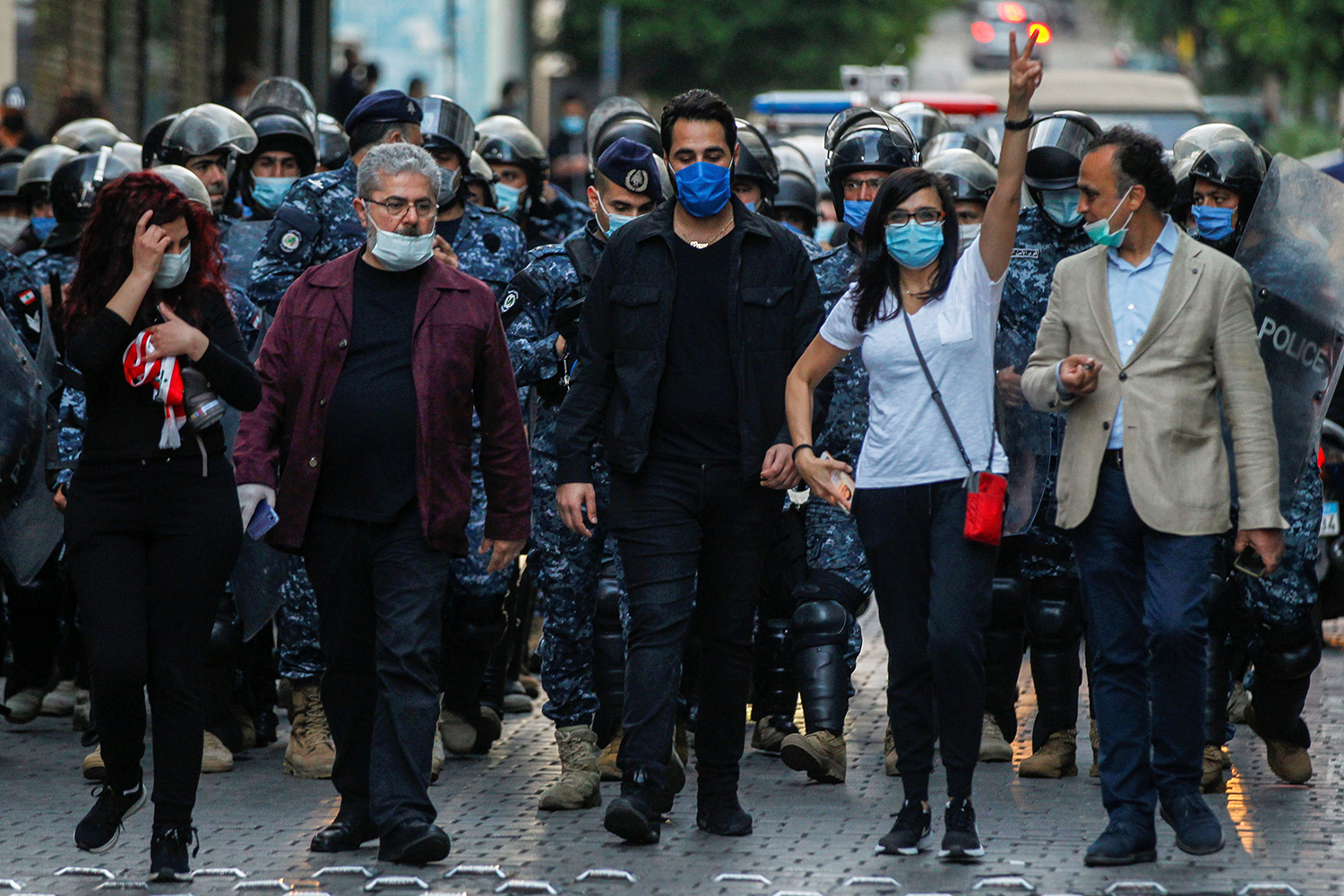 The photo shows a line of protestors wearing masks marching in front of a group of uniformed officers wearing riot gear. one woman is holding her hand in the air to make a victory/peace sign.