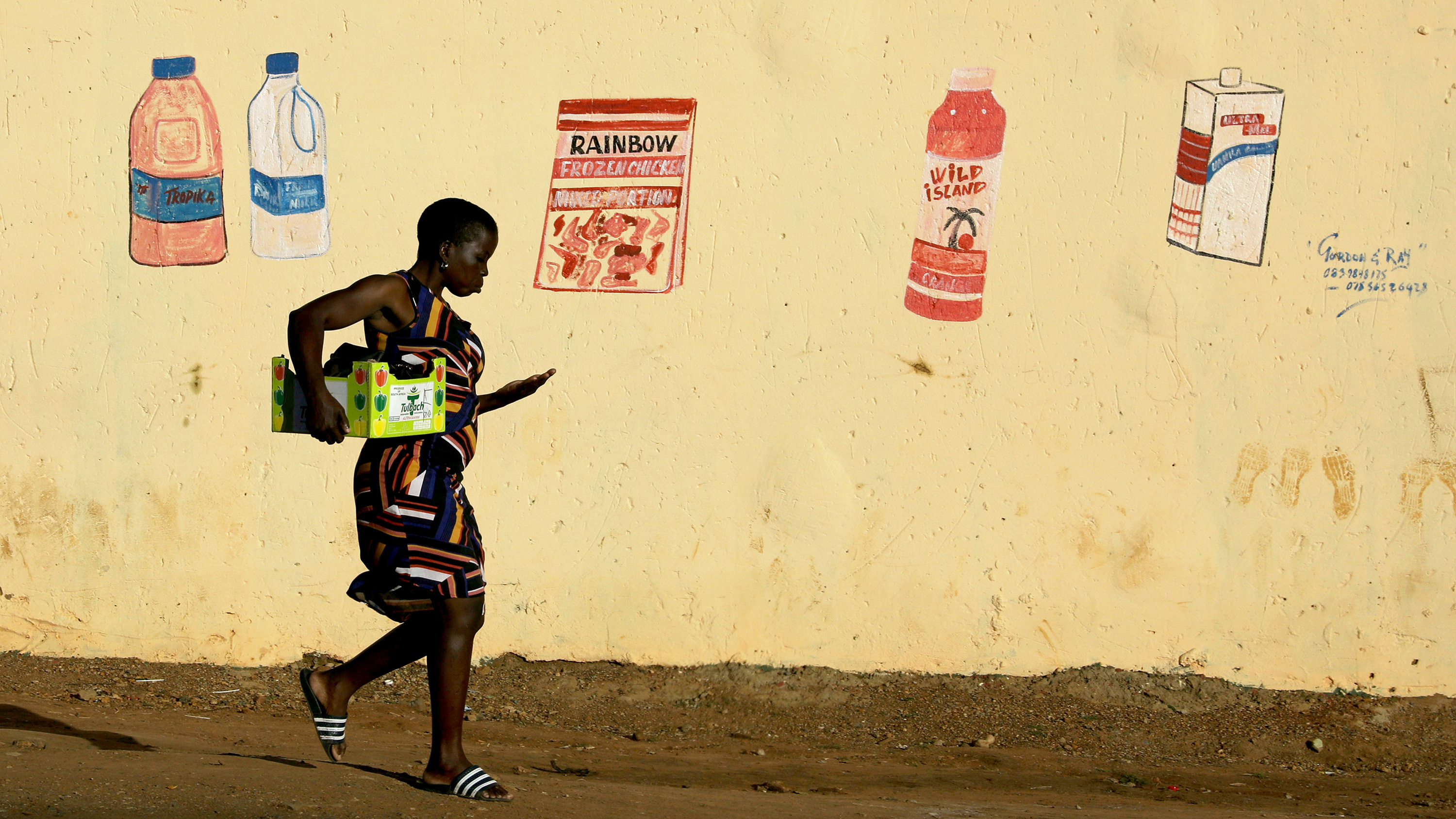 This is a striking photo of a woman carrying a box tucked under one arm and counting coins in the other walking in front of a butter yellow wall with faded, hand-drawn grocery items.