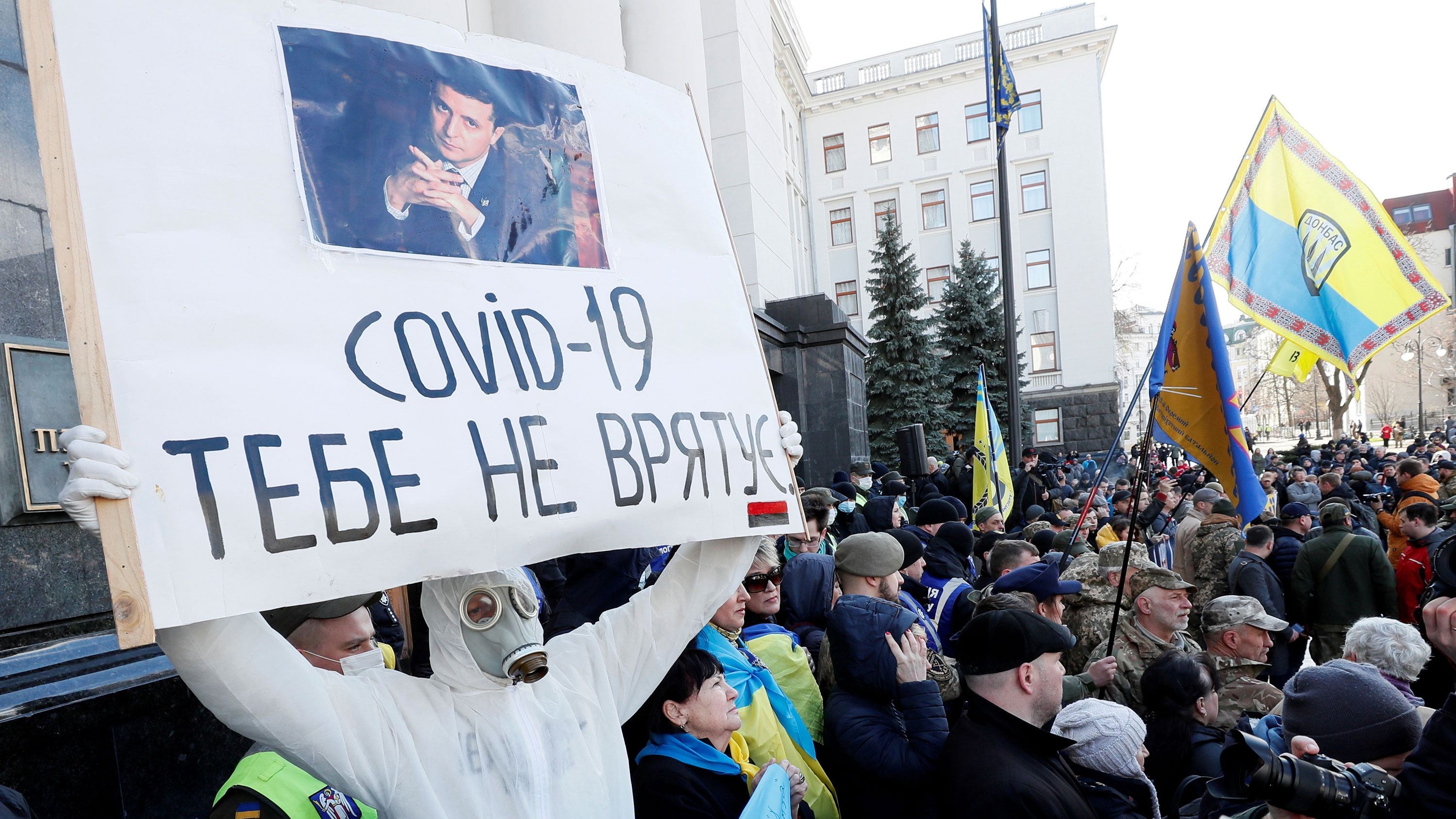 The photo shows a large protest with someone dressed in a suit resembling biocontainment gear holding the quoted sign.