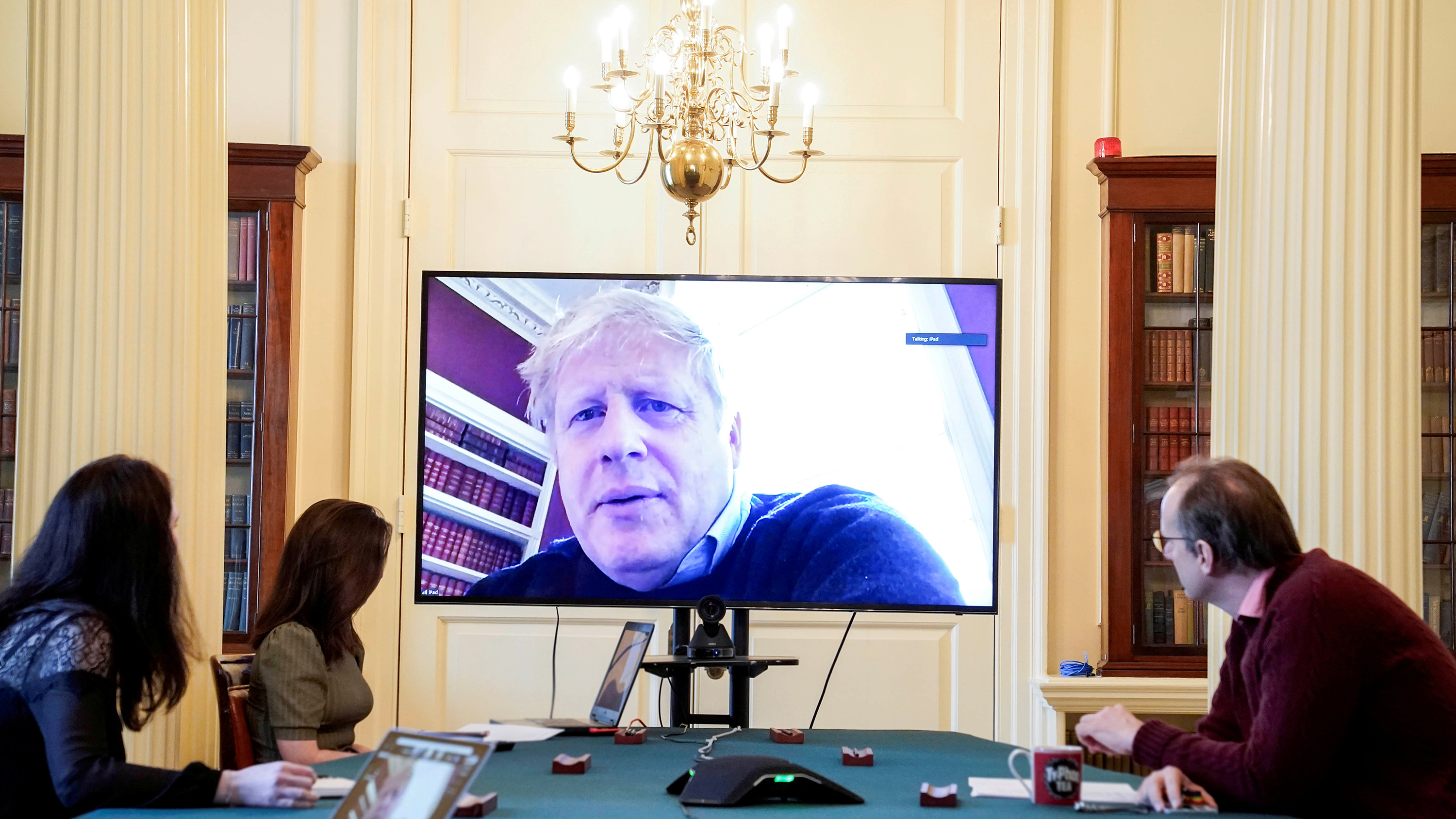 The photo is striking with people around a table and PM Johnson appearing on a monitor wheeled up to the head of the table.