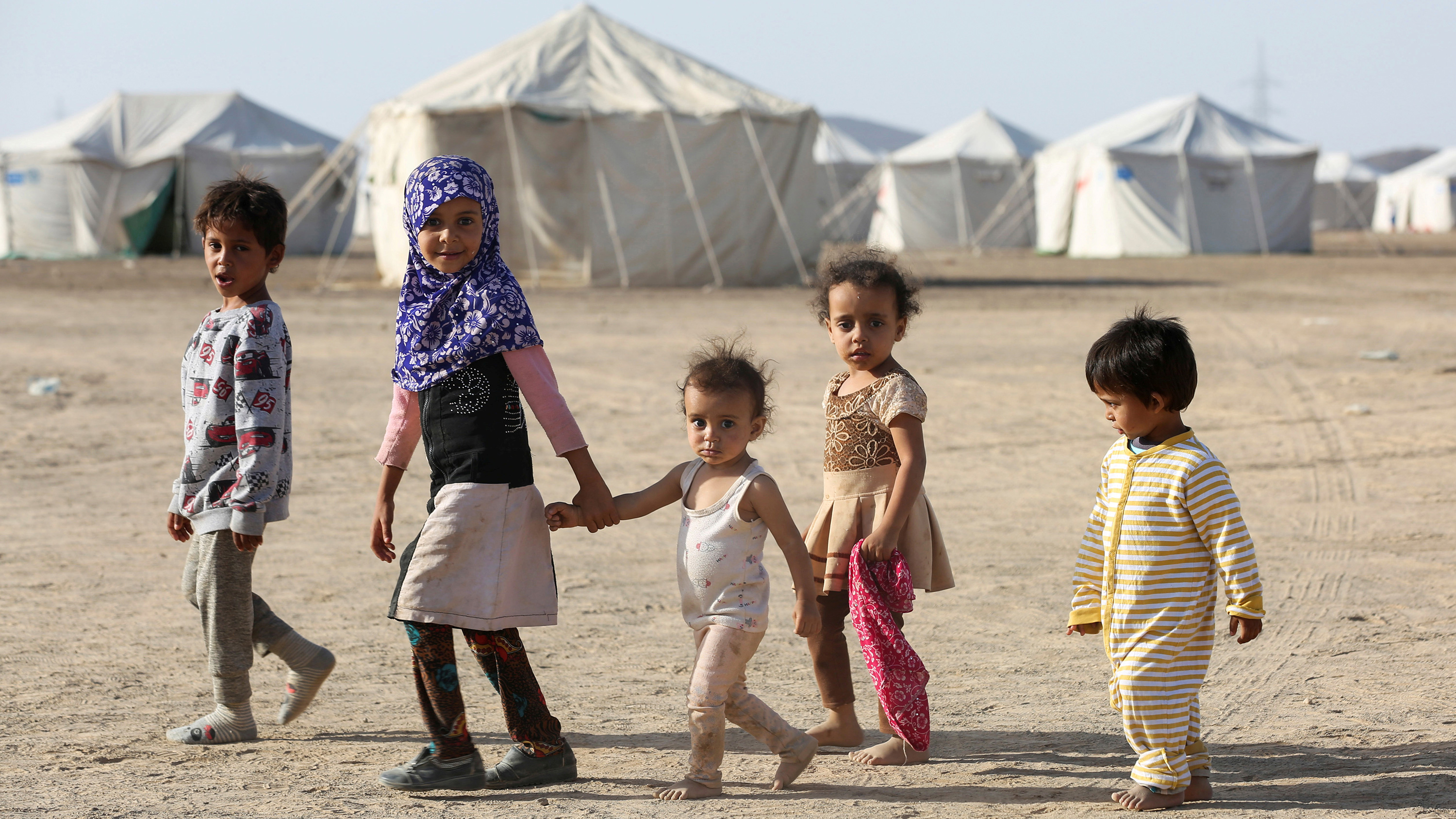 Picture shows a line of five children walking together in front of a camp filled with white tents and looking at the camera. All are very young, two who appear under eight years old, and three who appear under five.