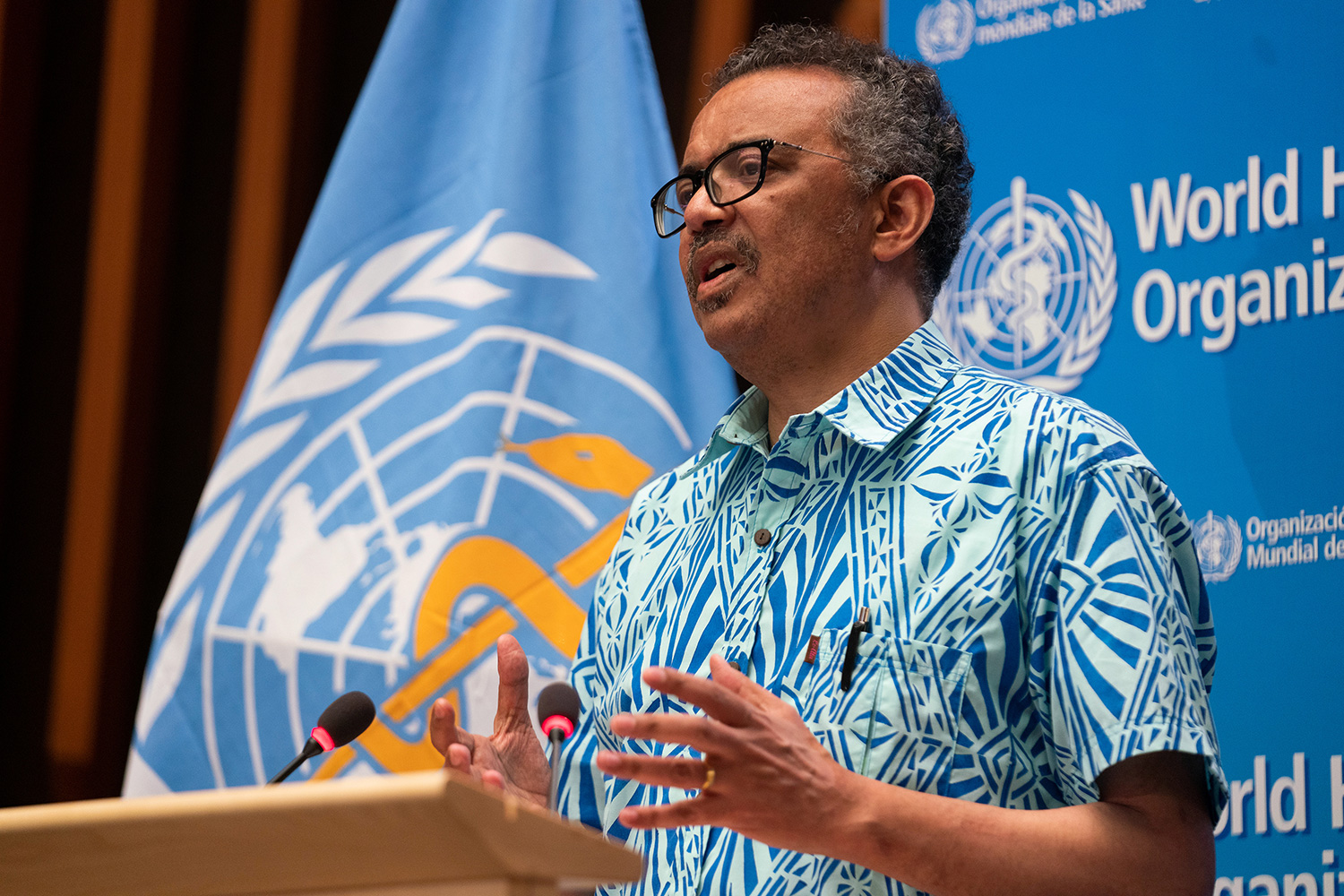 The WHO official is at a podium speaking, and surprisingly wearing a Hawaiian shirt.