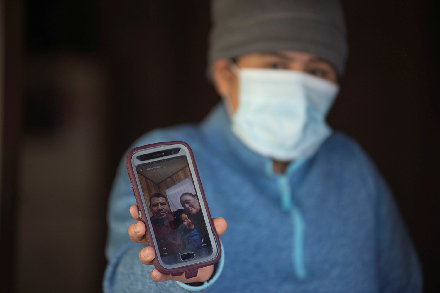 Picture shows Zoila holding a cell phone for the camera and wearing a mask. The photo on the phone shows the small family in happier times.