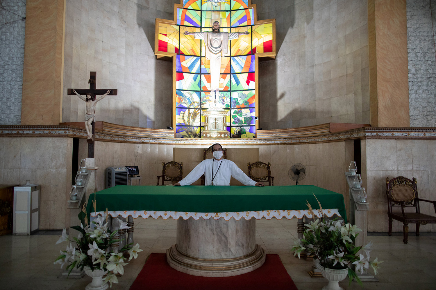 The photo shows the priest in front of a gorgeous stained glass window at the front of a church.