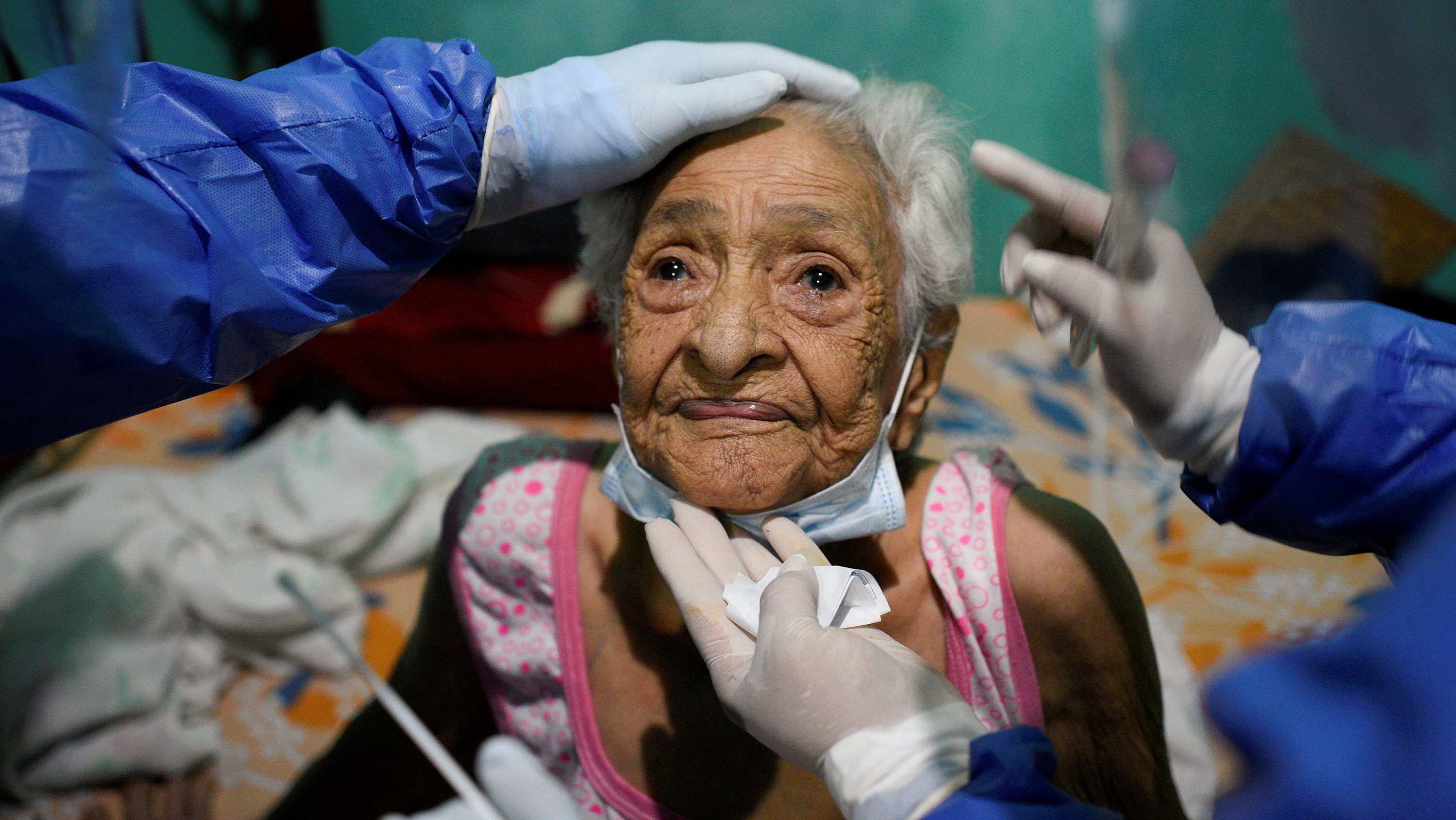 The photo shows an older women in the center of the frame with multiple glove-covered hands on her person.