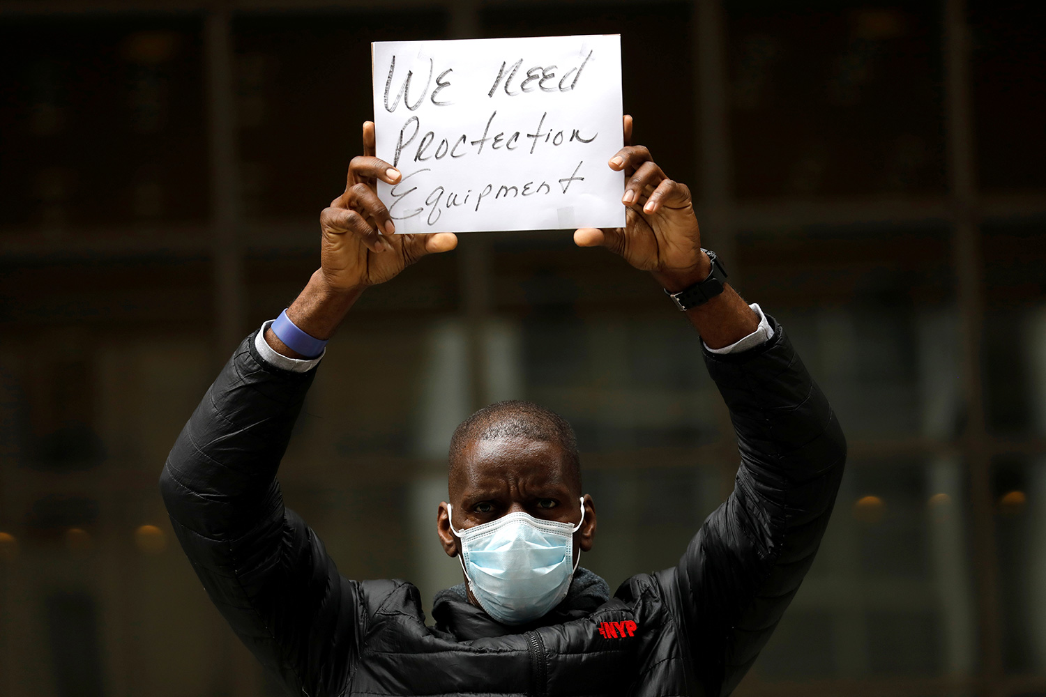 The image shows a man wearing a mask and holding a small sign above his head that says, "We need protection equipment."